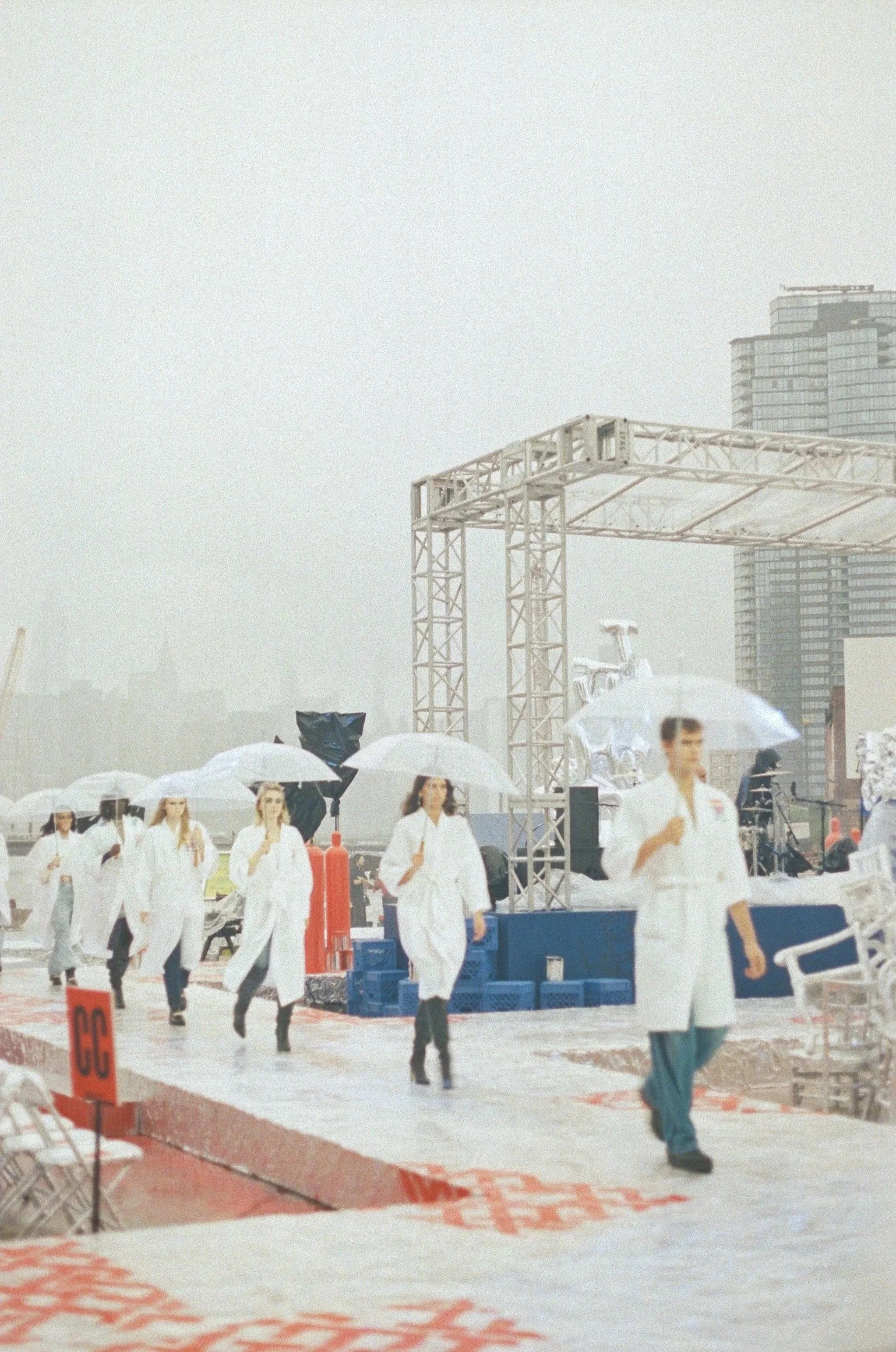 People dressed in white garments walking outdoors under umbrellas on a foggy day, with city buildings in the background, and construction equipment and scaffolding nearby.