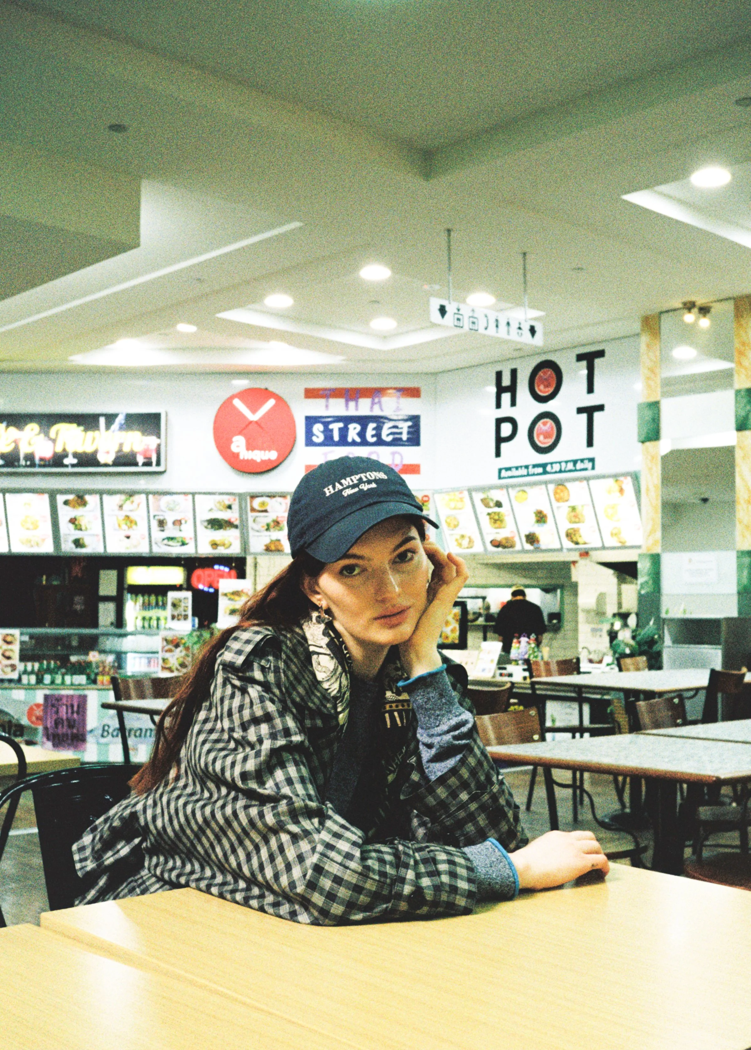 A young woman with long brown hair, wearing a black cap and checkered jacket, sitting at a table in a food court, resting her head on her hand and looking at the camera.