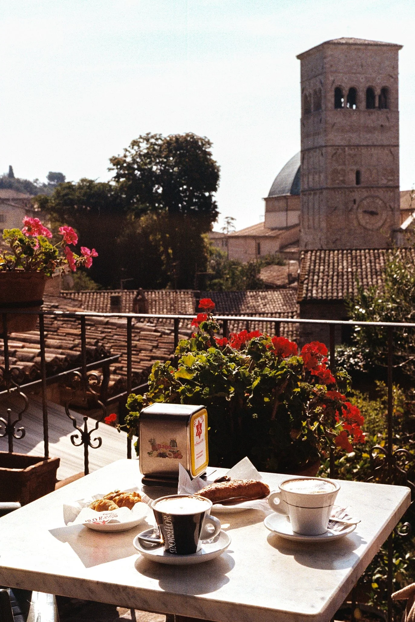 Outdoor cafe table with coffee, a pastry, and a flowerpot with pink and red flowers, overlooking rooftops and a historic stone bell tower in the background.