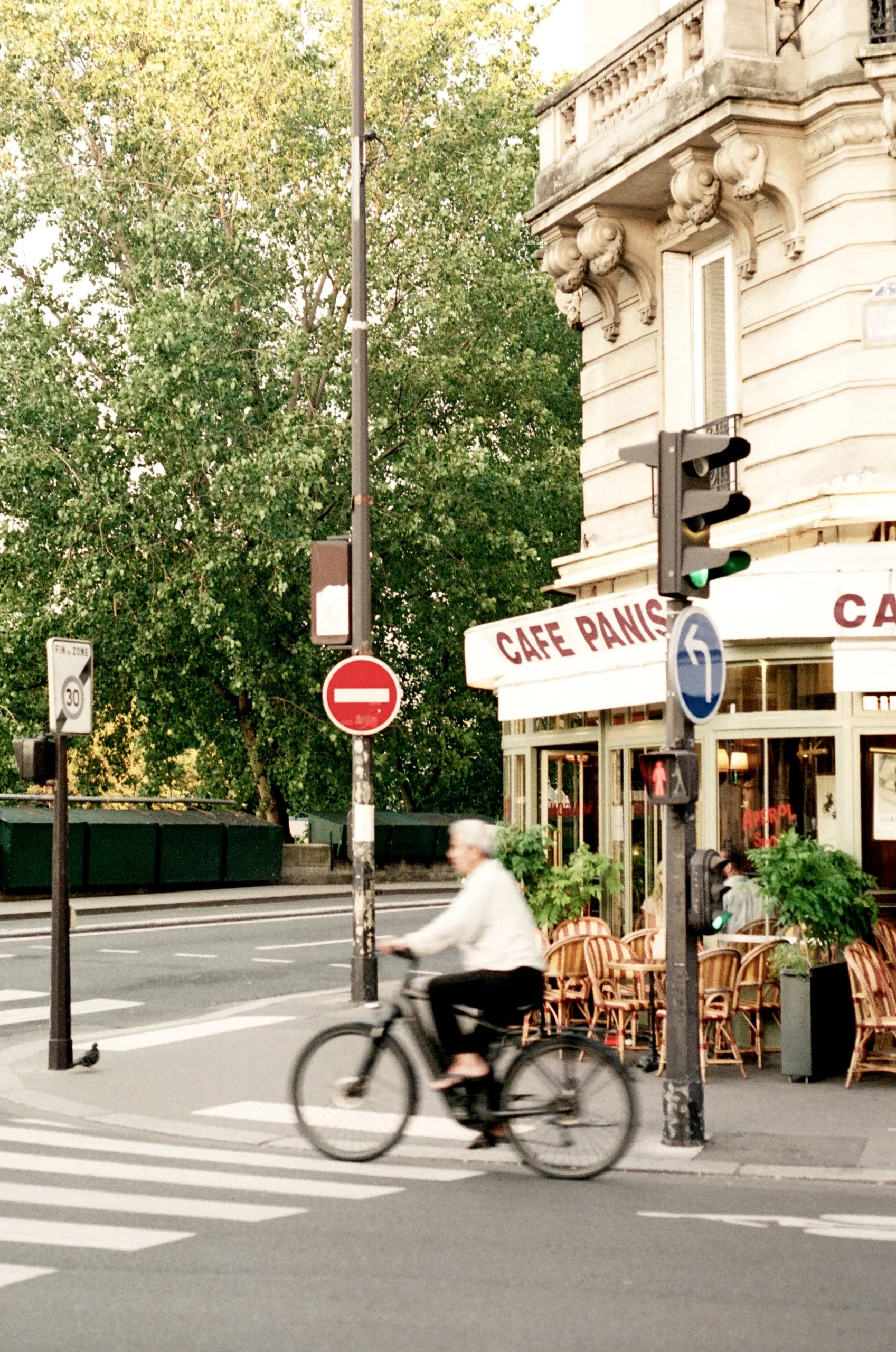 A person riding a bicycle past a cafe on a city street, with street signs, traffic lights, and greenery in the background.