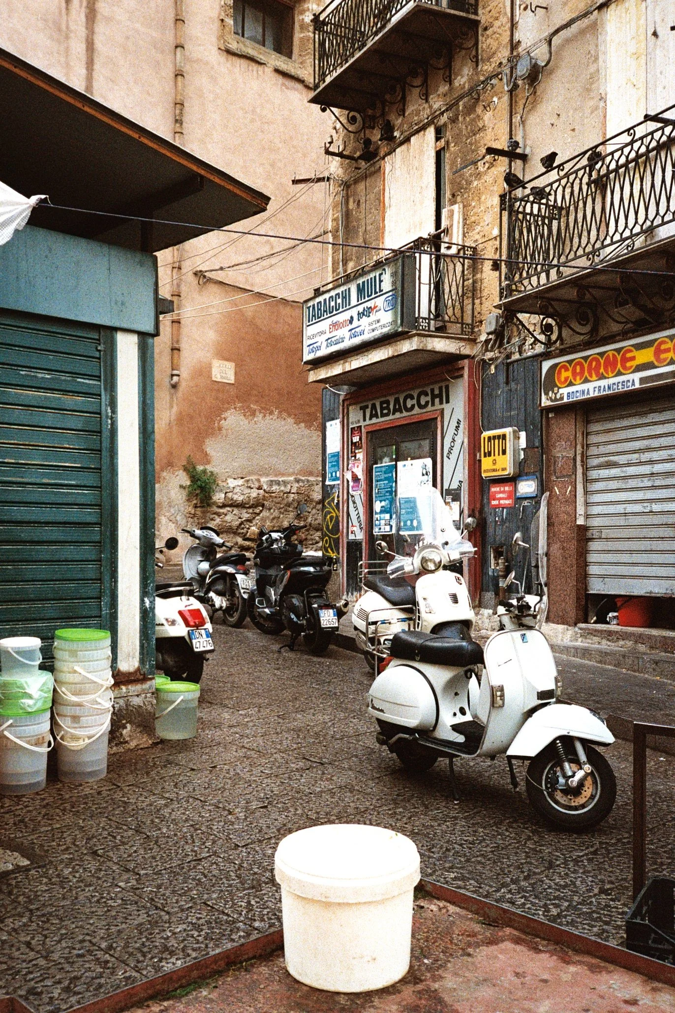 A narrow street scene with parked scooters and motorcycles, a storefront with a sign reading 'Tabacchi', and surrounding buildings with balconies and electronic shops.