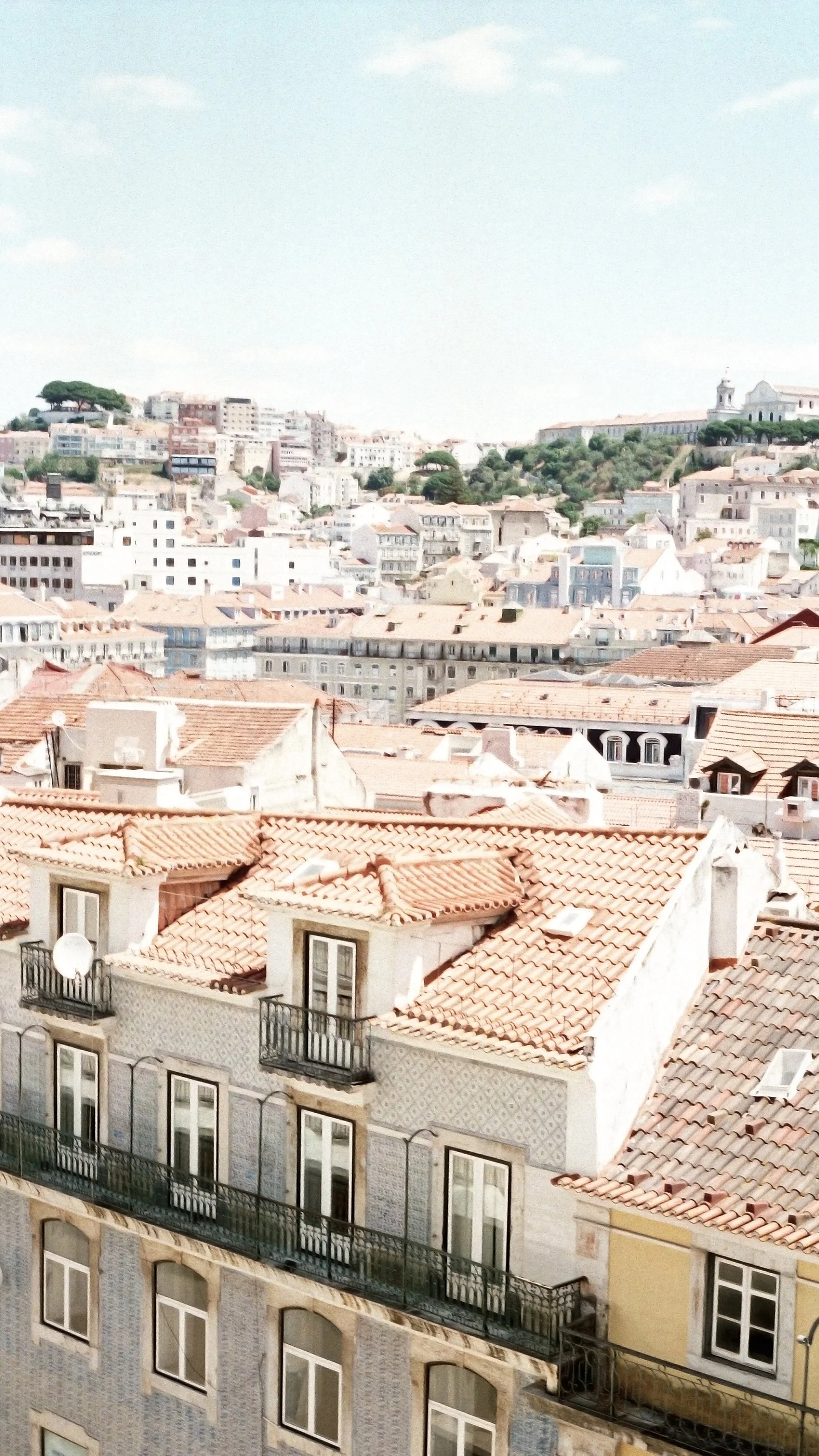 View of the rooftops and buildings of a hilly city, with a mix of older and modern architecture, under a partly cloudy sky.