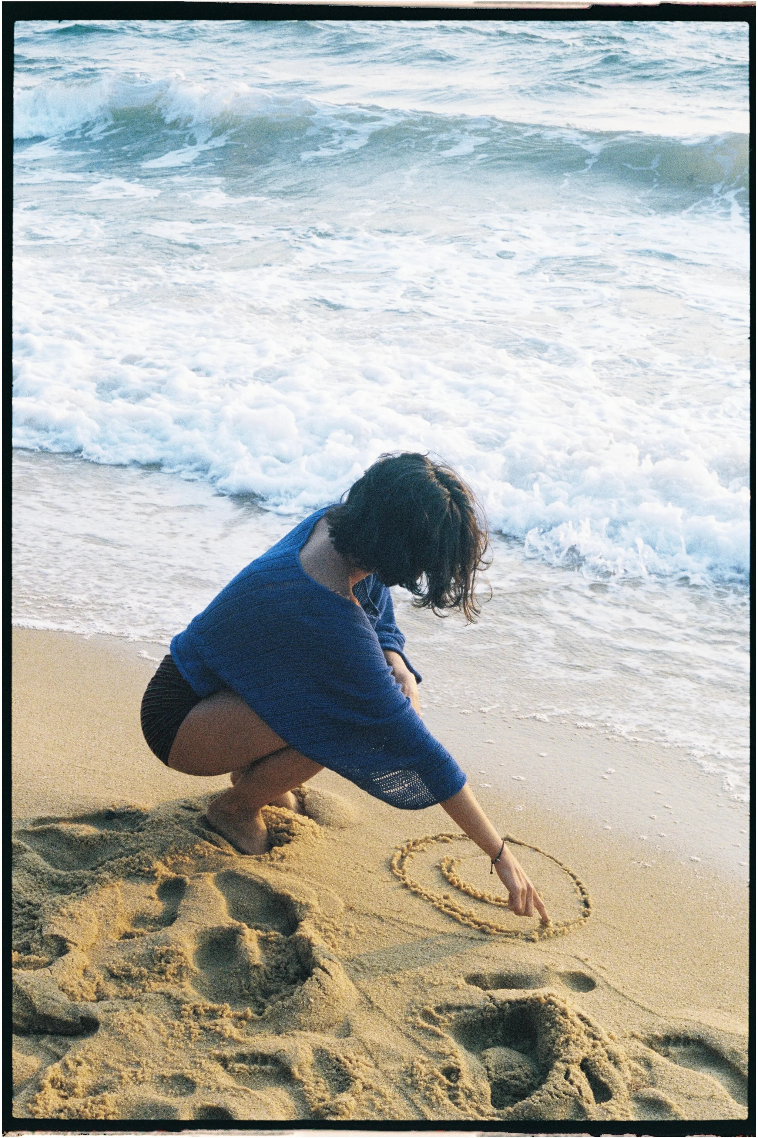 A person with dark hair, crouching on a sandy beach near the water's edge, drawing a heart shape in the sand with a stick.