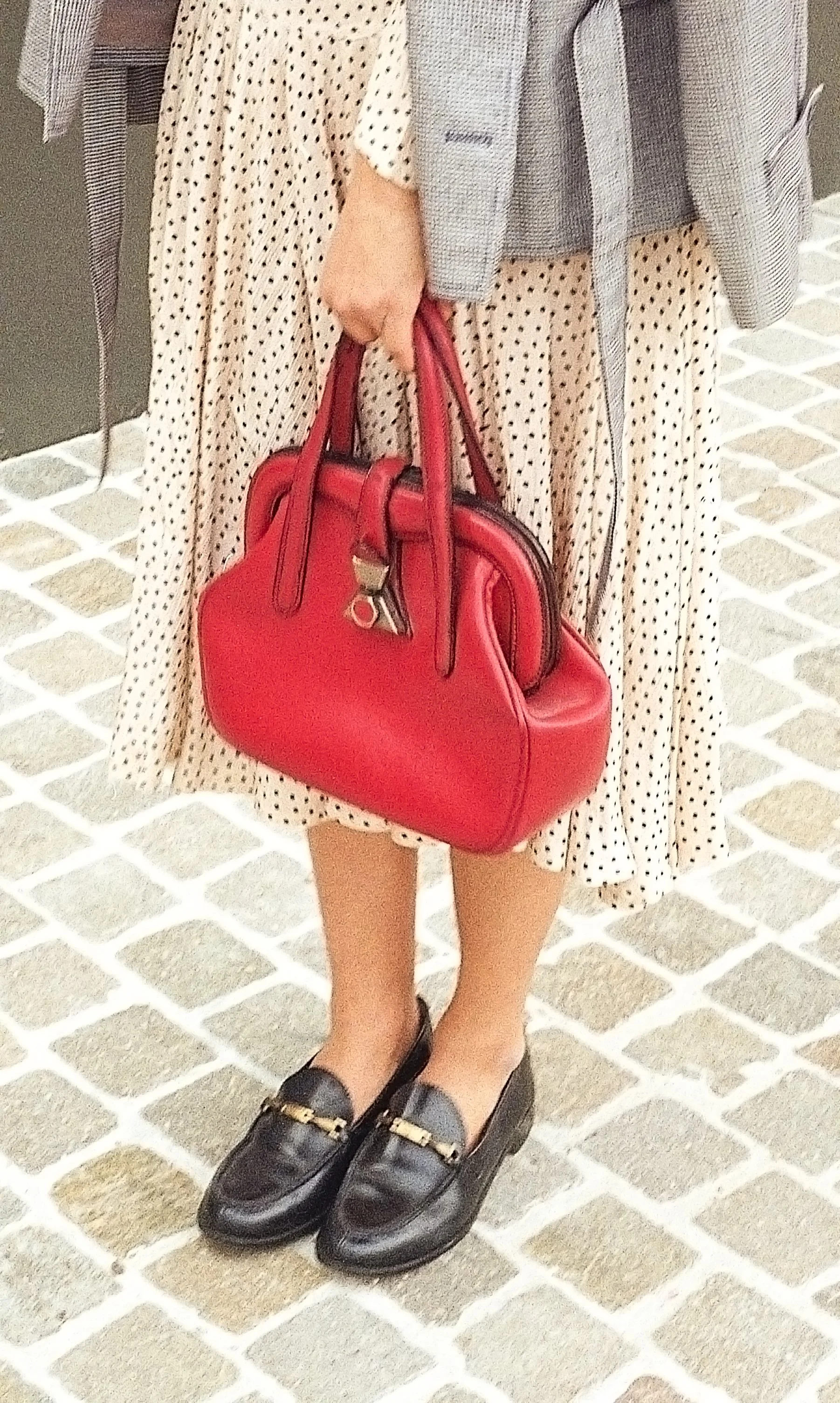 A woman holding a red handbag, wearing a polka dot dress, black loafers with gold accents, and a gray jacket.