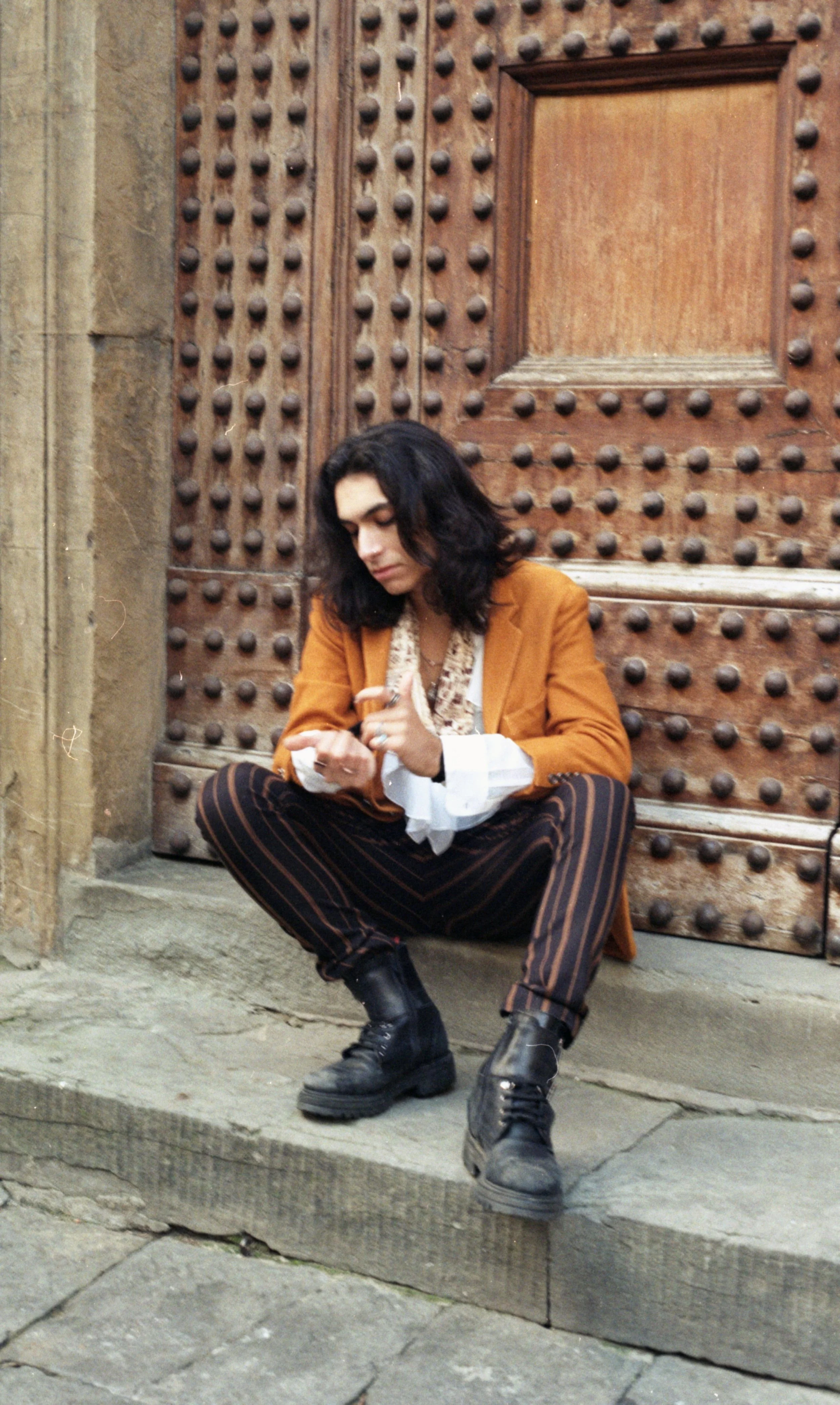 A woman with black hair wearing a brown blazer, striped pants, and black combat boots sitting on stone steps in front of a wooden door with metal studs, looking at her phone.