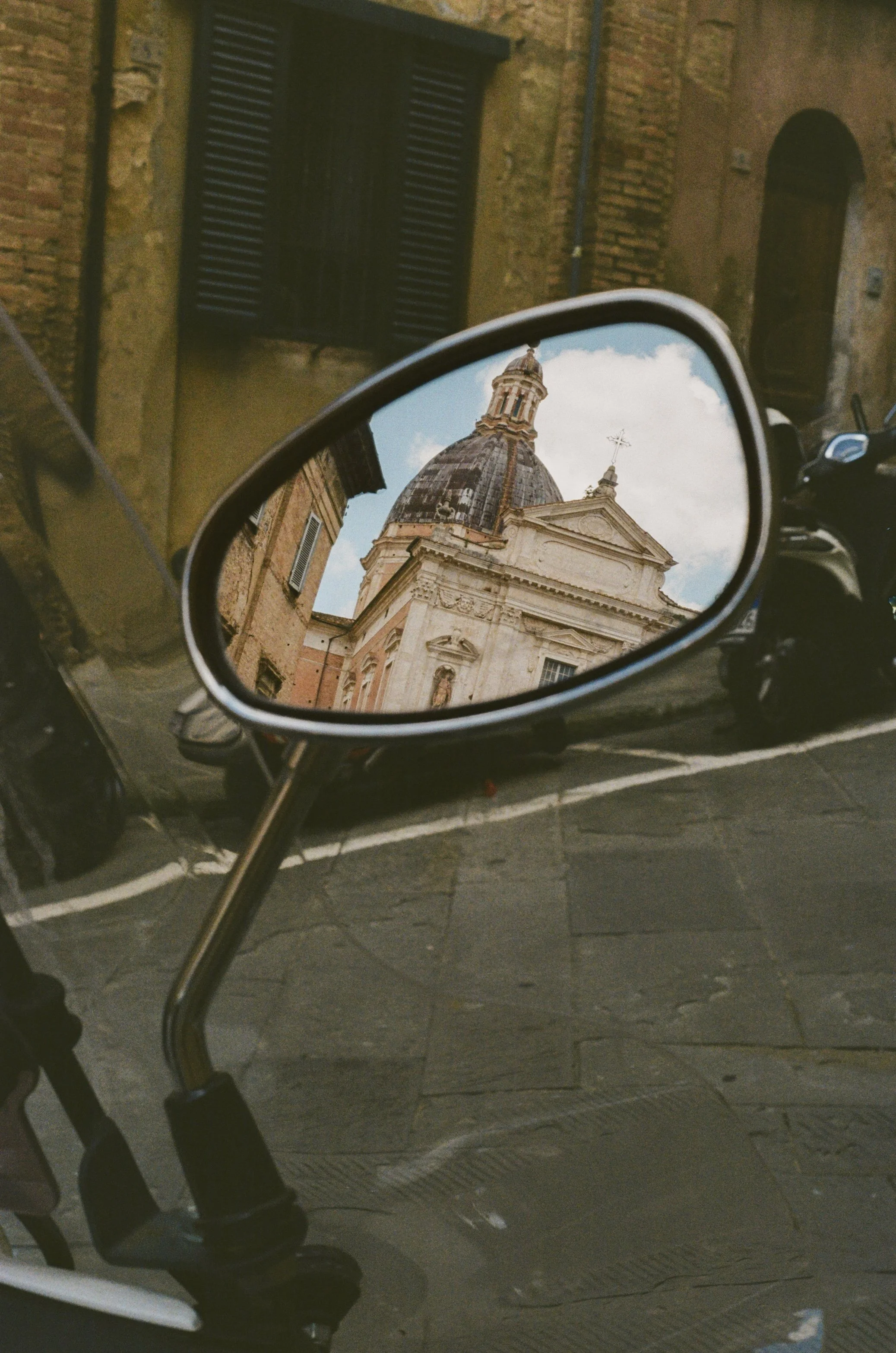Reflection of a historic church with a domed roof and detailed facade in a motorcycle's side mirror on a cobblestone street.