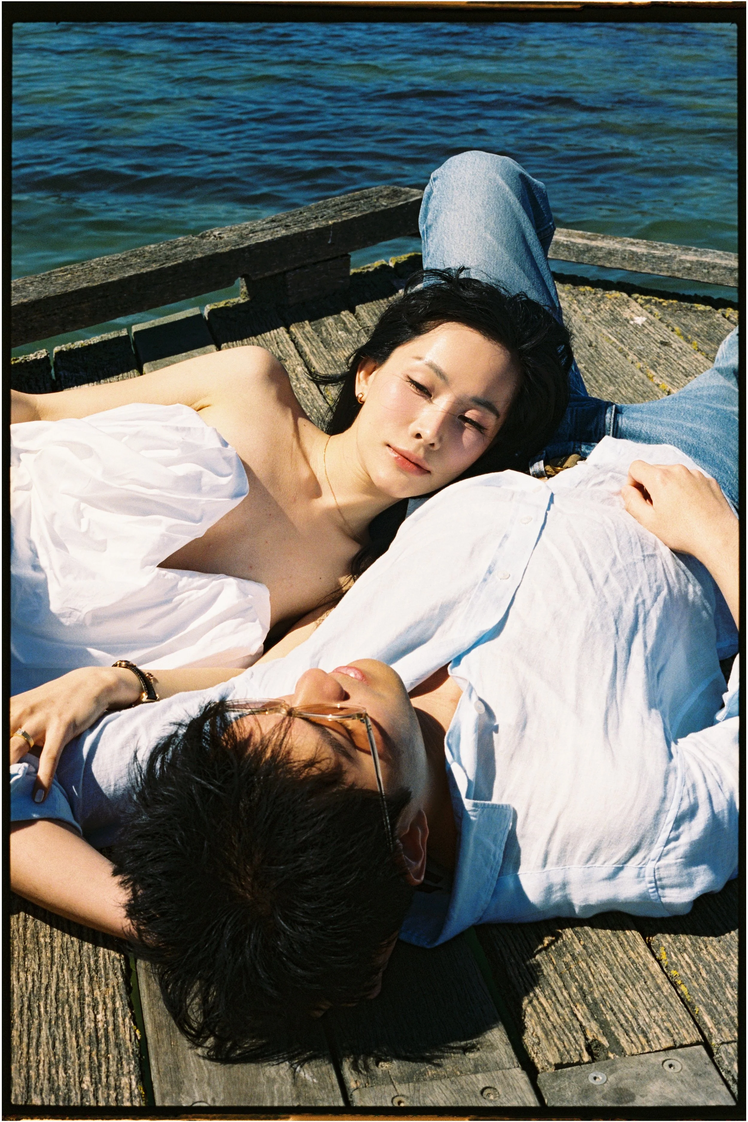 A young couple lying on a wooden dock near water, relaxed and enjoying the sunlight.
