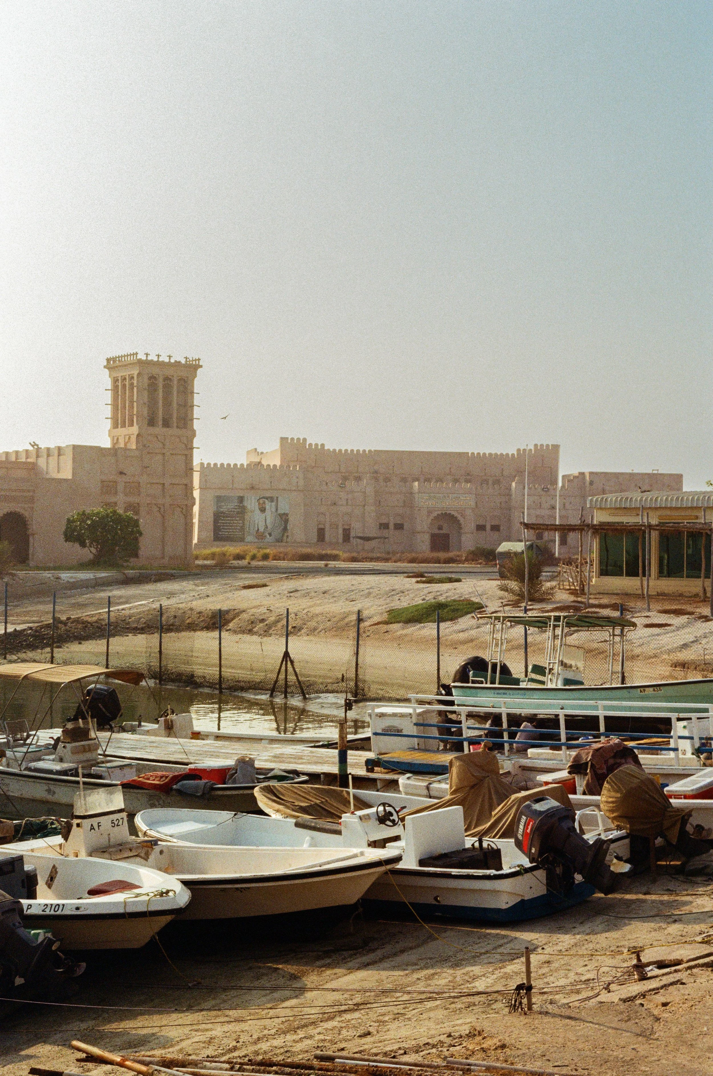 A harbor scene with several small boats docked on the beach, in front of a sandy area and a historic fort with a tower and walls in the background, under a clear blue sky.