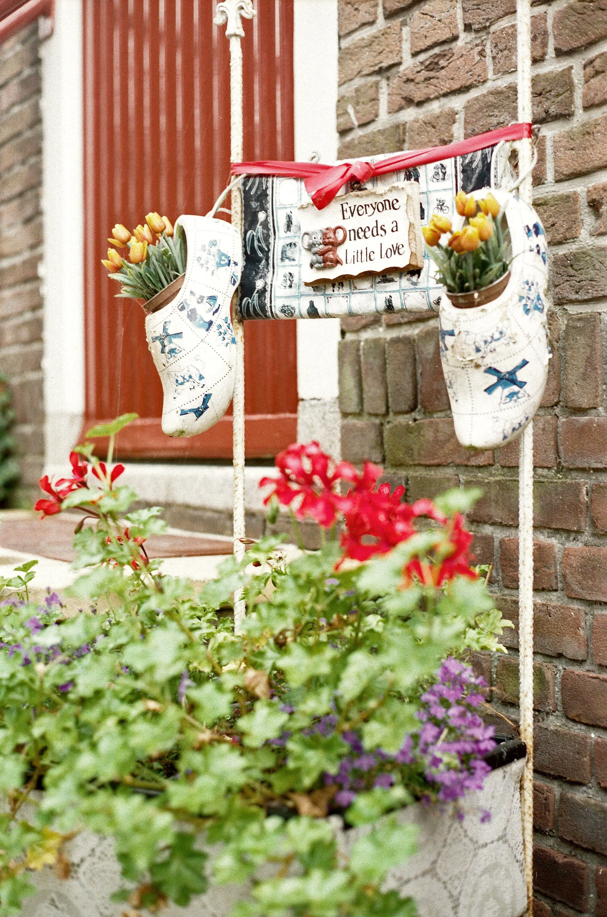 Decorative hanging shoes with tulips, a sign reading 'Everyone needs a little love,' and colorful flowers in a container on a brick porch wall.