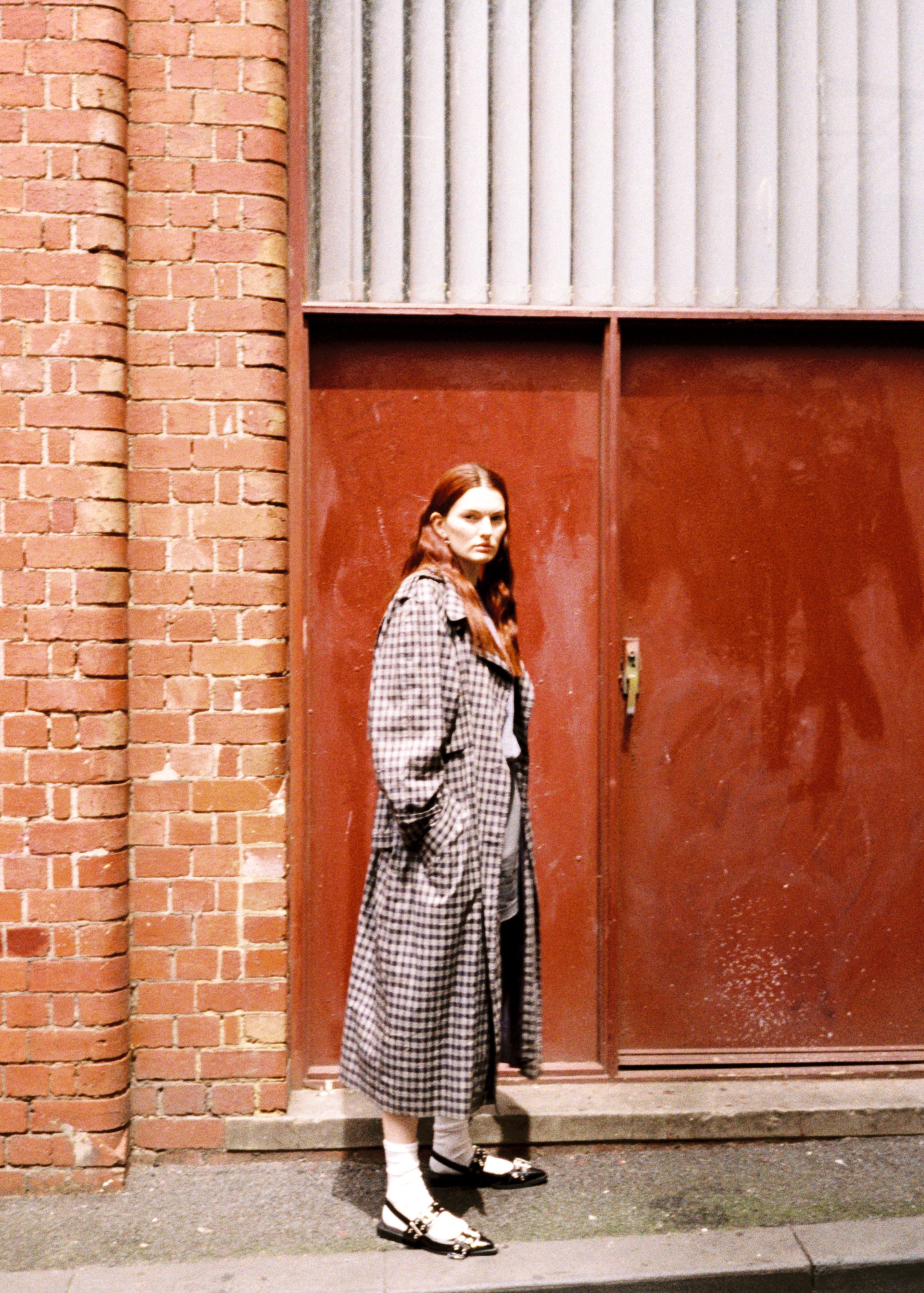 A young woman with long red hair standing in front of a red metal door with a brick wall on the left side. She is wearing a checkered coat, gray pants, white socks, and black shoes.