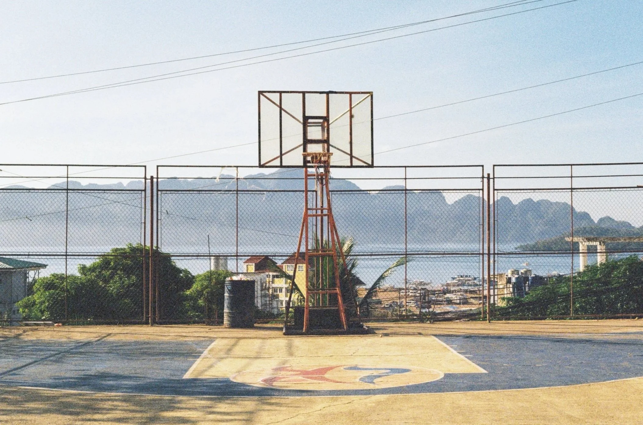 An outdoor basketball court with a single hoop, surrounded by a chain-link fence, with buildings and mountains in the background under a cloudy sky.