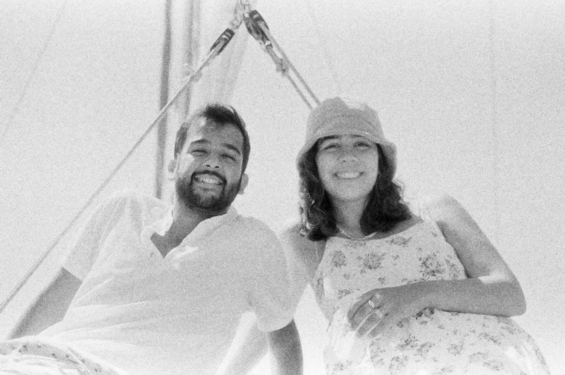A black and white photo of a smiling man and woman outdoors, with a pole and flag in the background. The man has a beard and is wearing a light-colored shirt, while the woman has a hat and a floral dress.