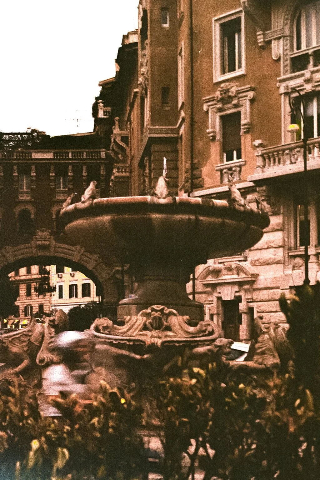 A vintage-style photograph showing a large fountain with ornate sculptures of sea creatures, situated in front of historic European buildings with balconies and decorative windows.