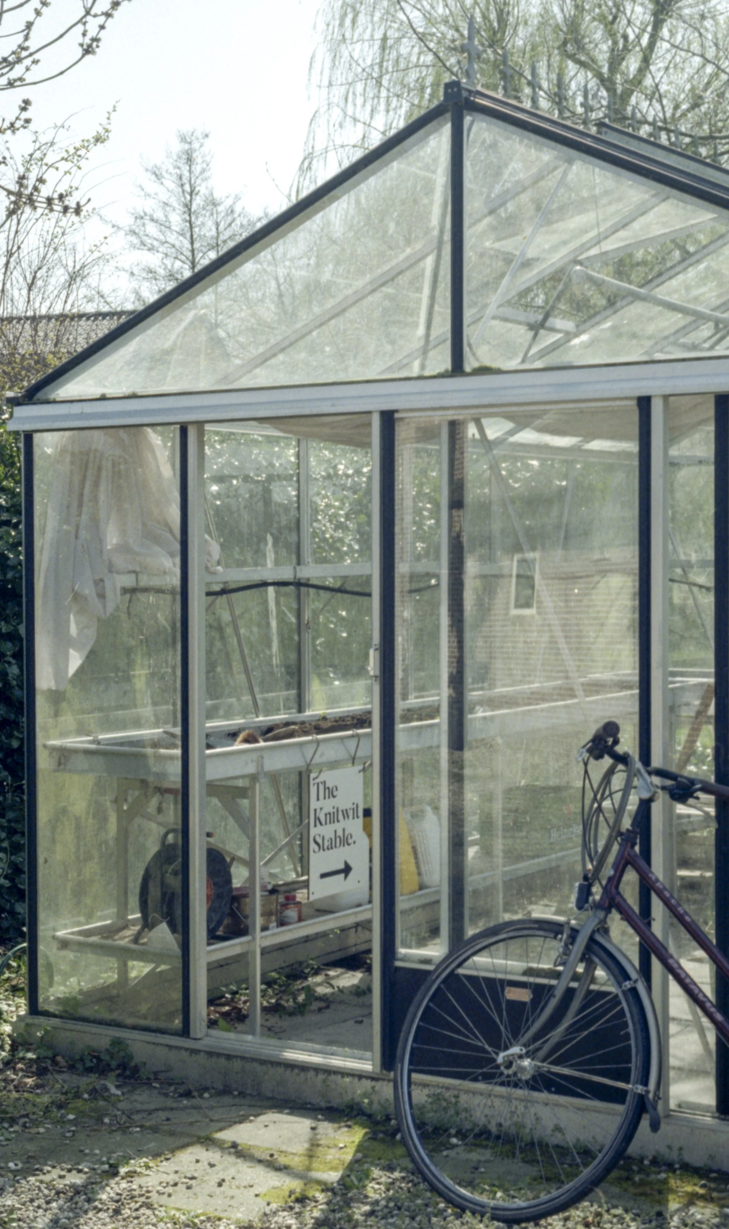 A small greenhouse with a sign inside that reads "The Knitwit Stable" and points to the right. There is a bicycle parked outside the greenhouse. Through the glass, some items and a person are visible inside.