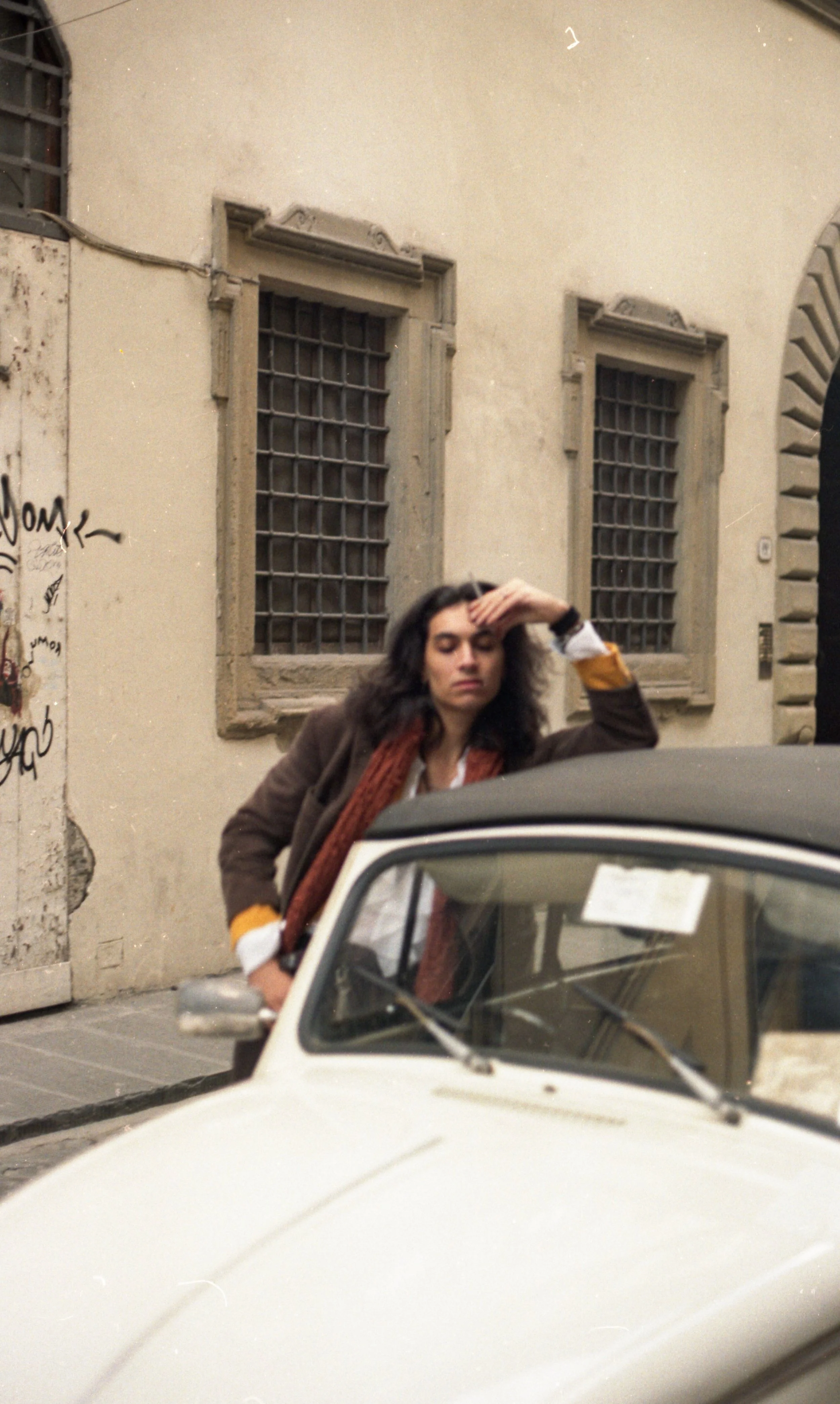 A woman with dark curly hair, wearing a brown coat and a red scarf, is standing behind a white vintage car on a city street. She has her right hand on her forehead, appearing to be tired or stressed, and is looking downward. The background features a beige building with gritted windows and graffiti on a wall.