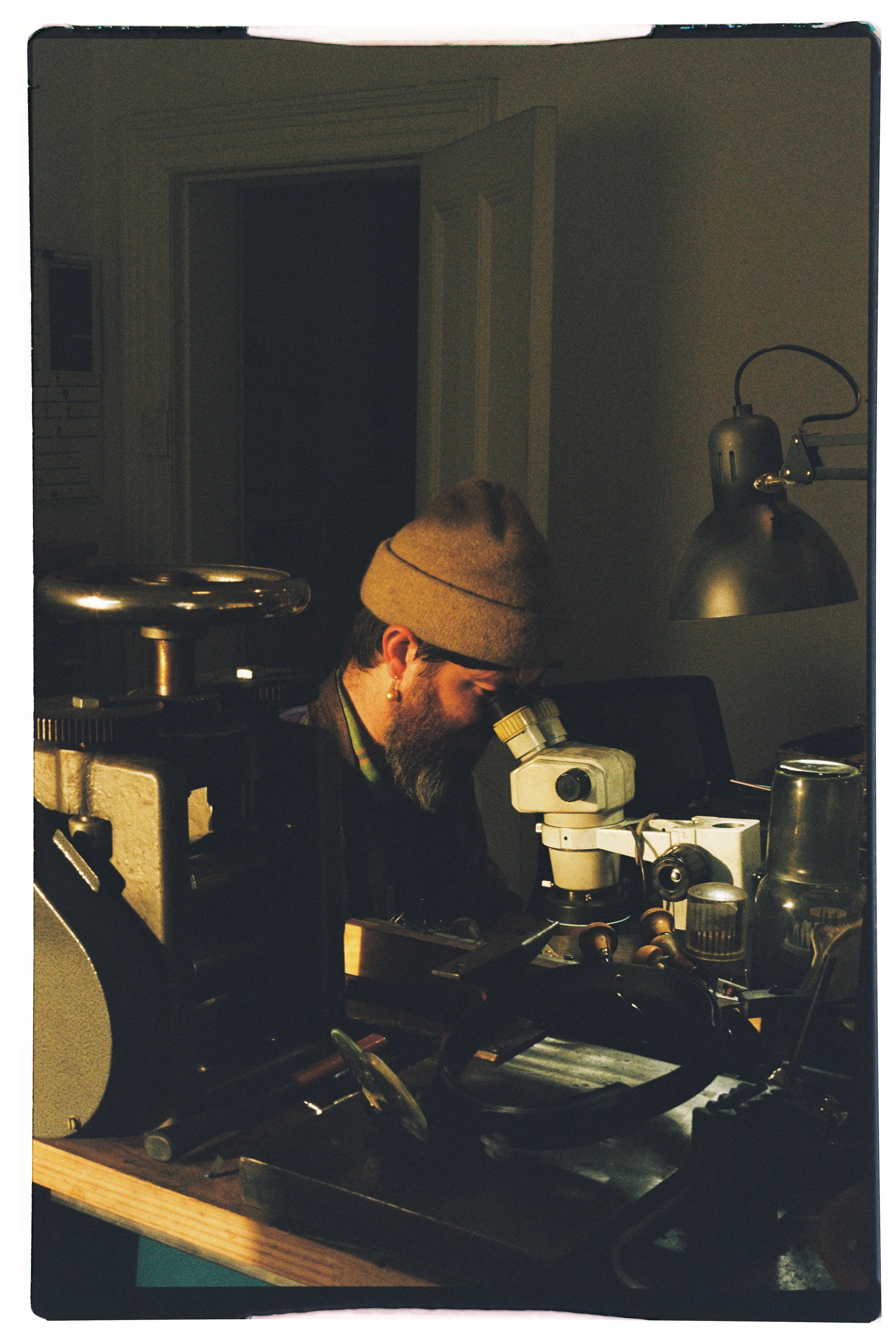 A man with a beard wearing a brown beanie, looking into a microscope in a dimly lit room, surrounded by scientific equipment.