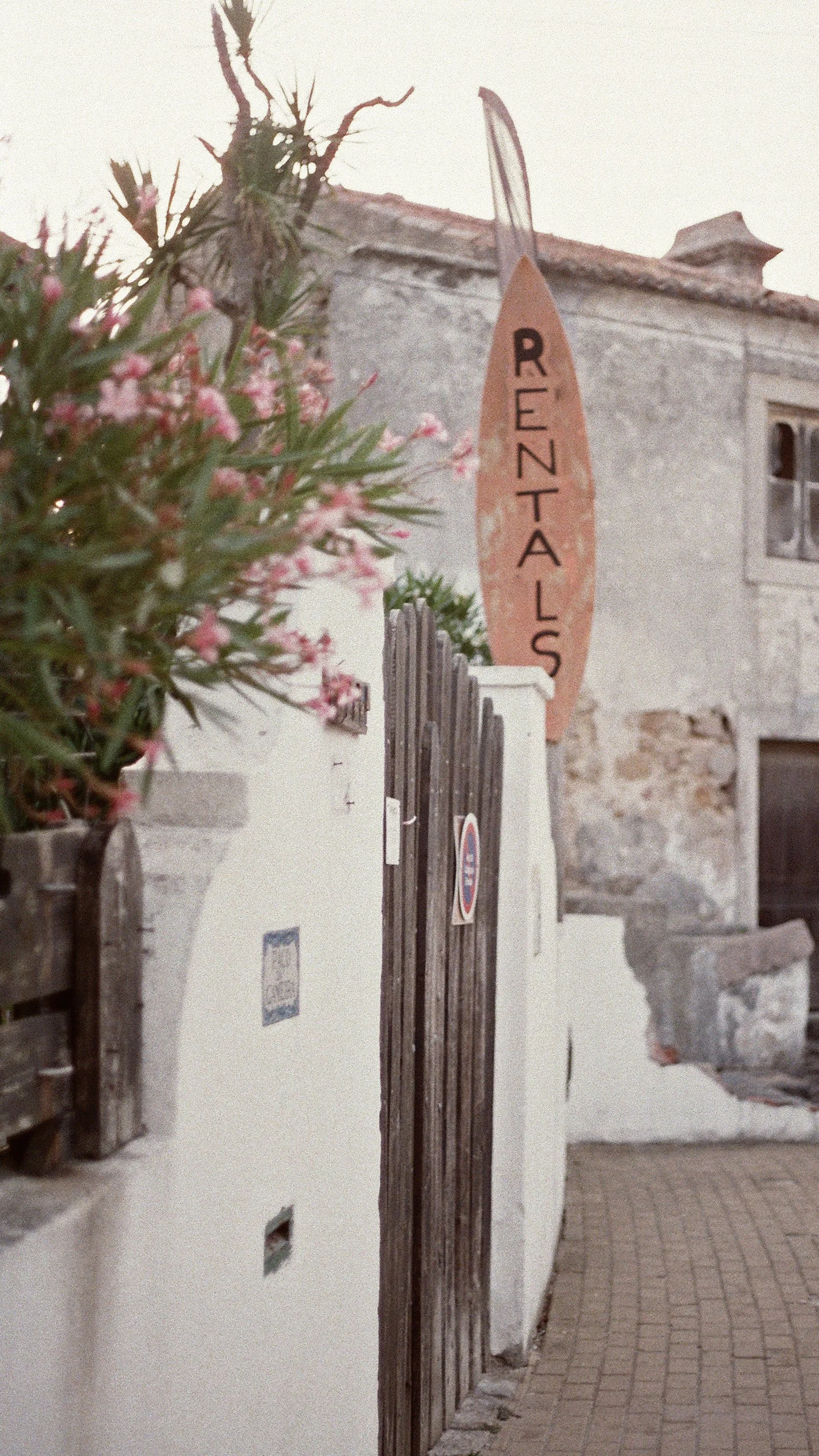 A wooden gate with a sign above that reads 'RESTAURANT'. The gate is on a cobblestone pathway next to a white wall with some small vent openings and some greenery with pink flowers in the foreground, and an old building with a small window and weathered exterior in the background.