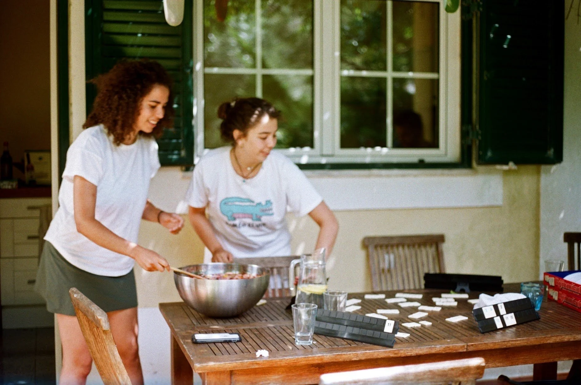 Two women preparing food together in a bright kitchen, with open window and outdoor greenery visible, on a wooden table with game pieces and a phone.