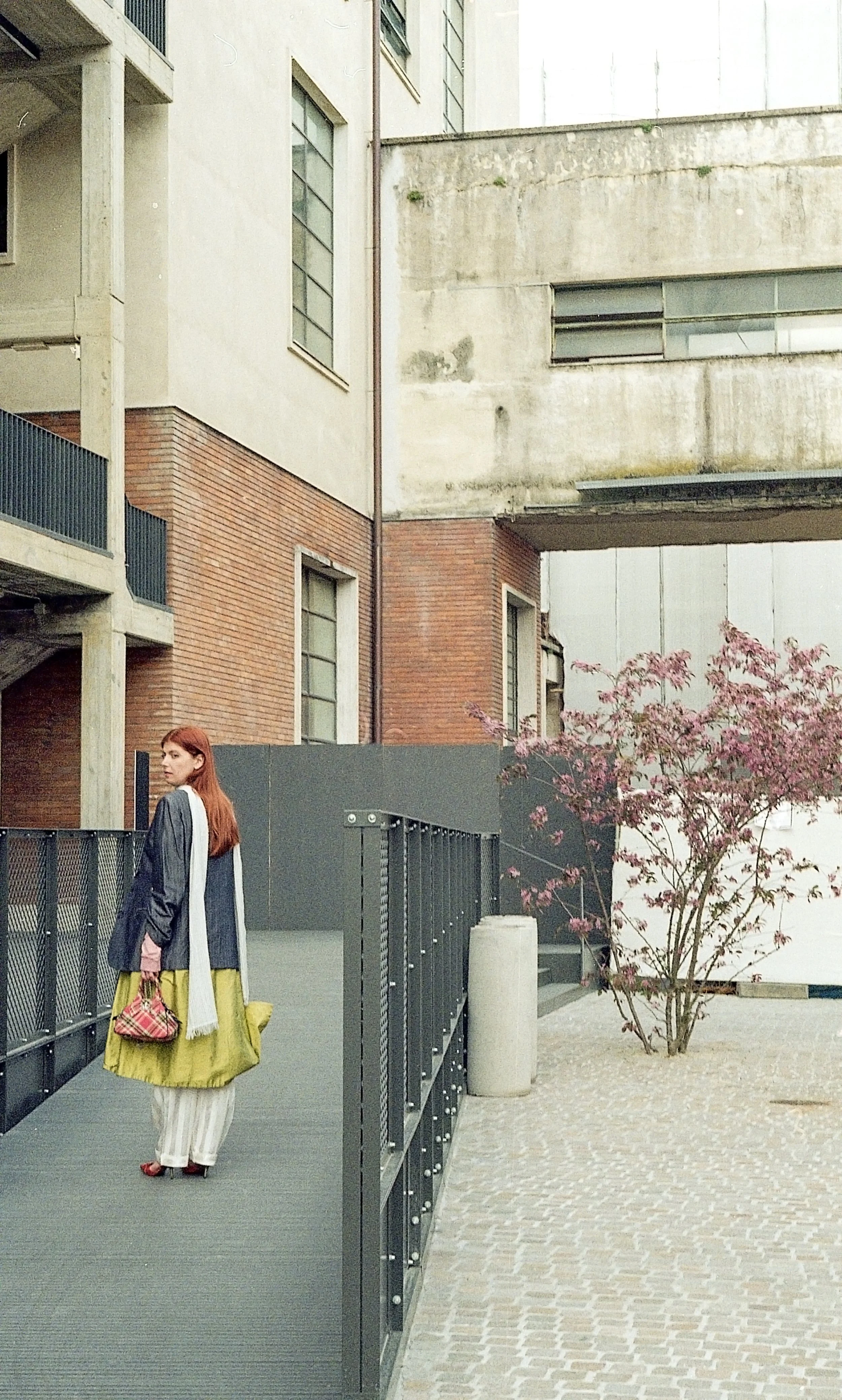 A woman with red hair wearing a dark jacket, a yellow skirt, and striped pants holds a small plaid purse. She stands on a modern outdoor walkway with black railing, next to a pink flowering tree and urban building structures.