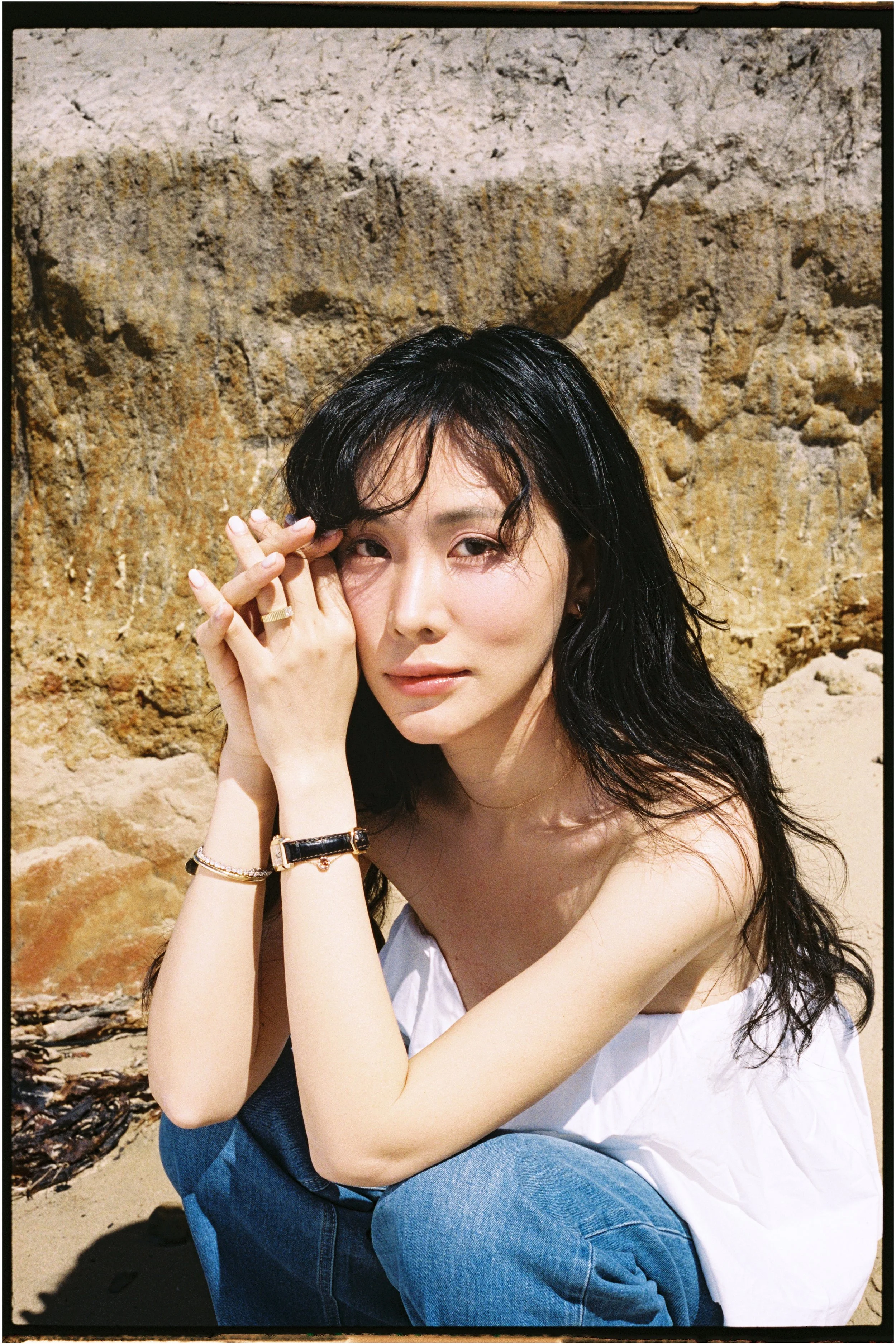 A woman with black wavy hair and wearing a white sleeveless top, sitting on the beach with a rocky background, looking at the camera with a relaxed expression.