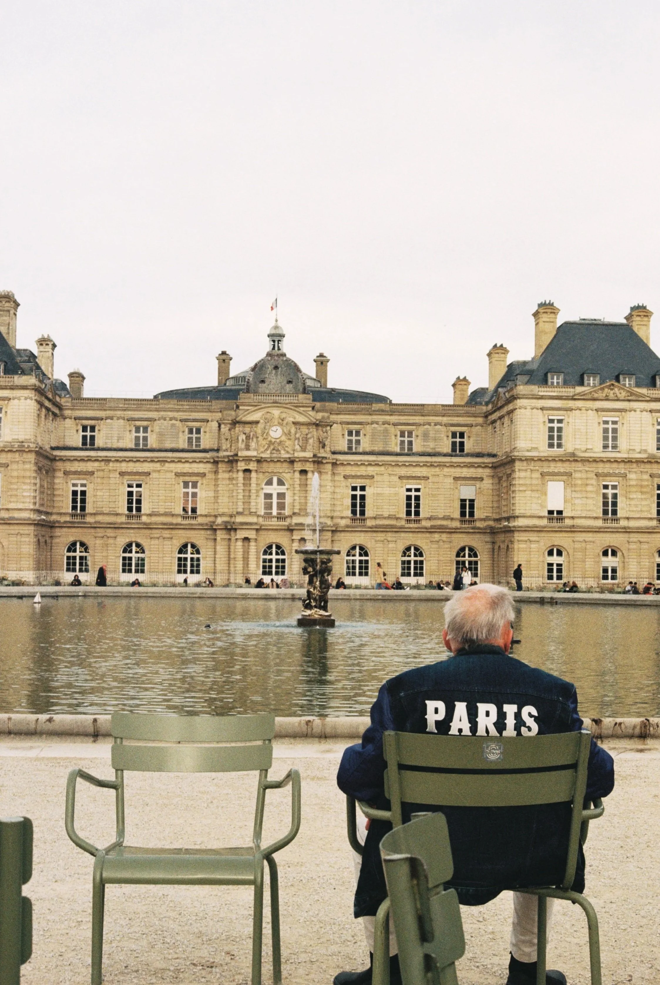 An elderly man with gray hair, wearing sunglasses and a denim jacket with 'PARIS' written on the back, sitting on a green chair in front of a fountain in a park with a large historic building in the background.