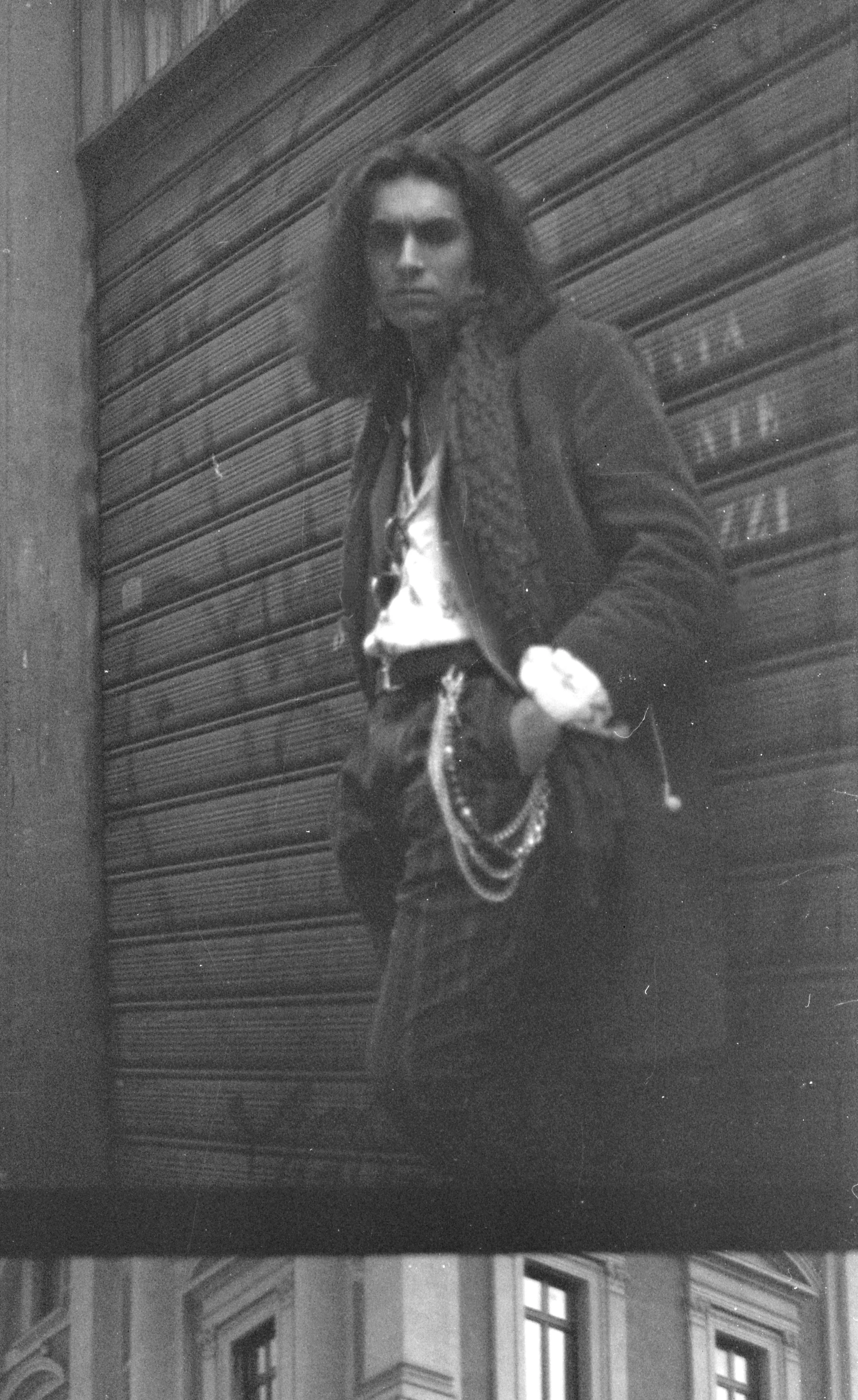 Black and white photo of a man with long curly hair standing against a closed metal shutter, wearing a jacket with a chain and a scarf.