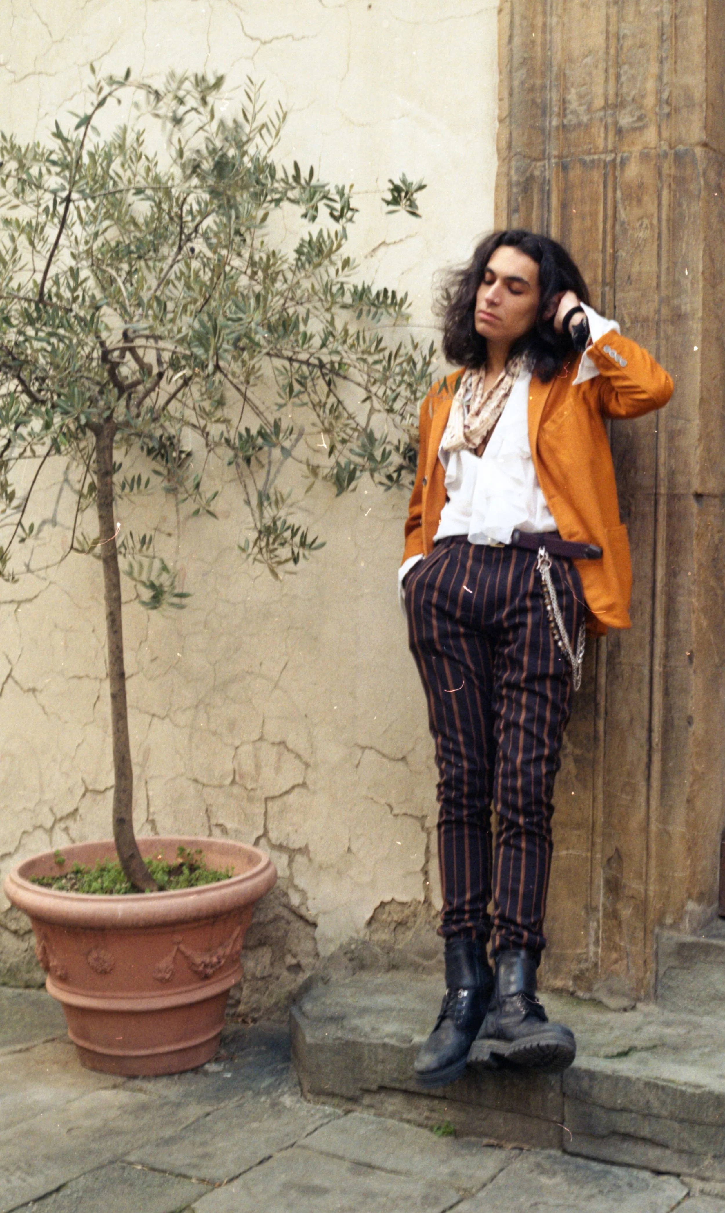 A person with long dark curly hair leaning against a textured wooden wall, wearing a mustard blazer, white shirt, patterned scarf, striped pants with chains, and black boots, standing next to a potted plant with an olive tree, on a stone pavement.