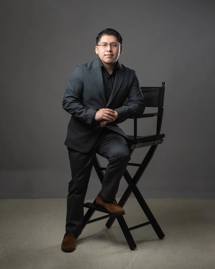 Man wearing black suit and glasses, leaning on a director's chair against a neutral gray background.