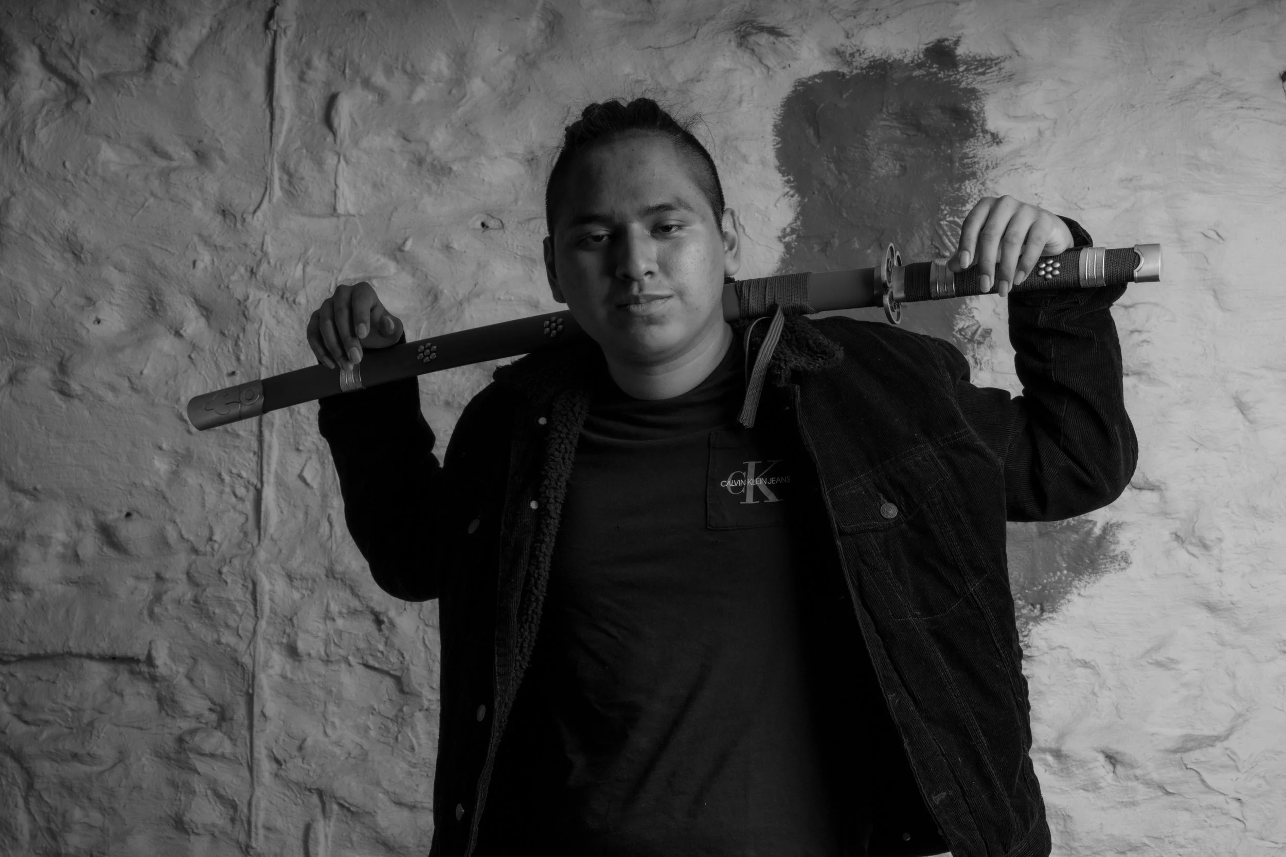 A young man holding a stick over his shoulders, standing against a textured wall in black and white.