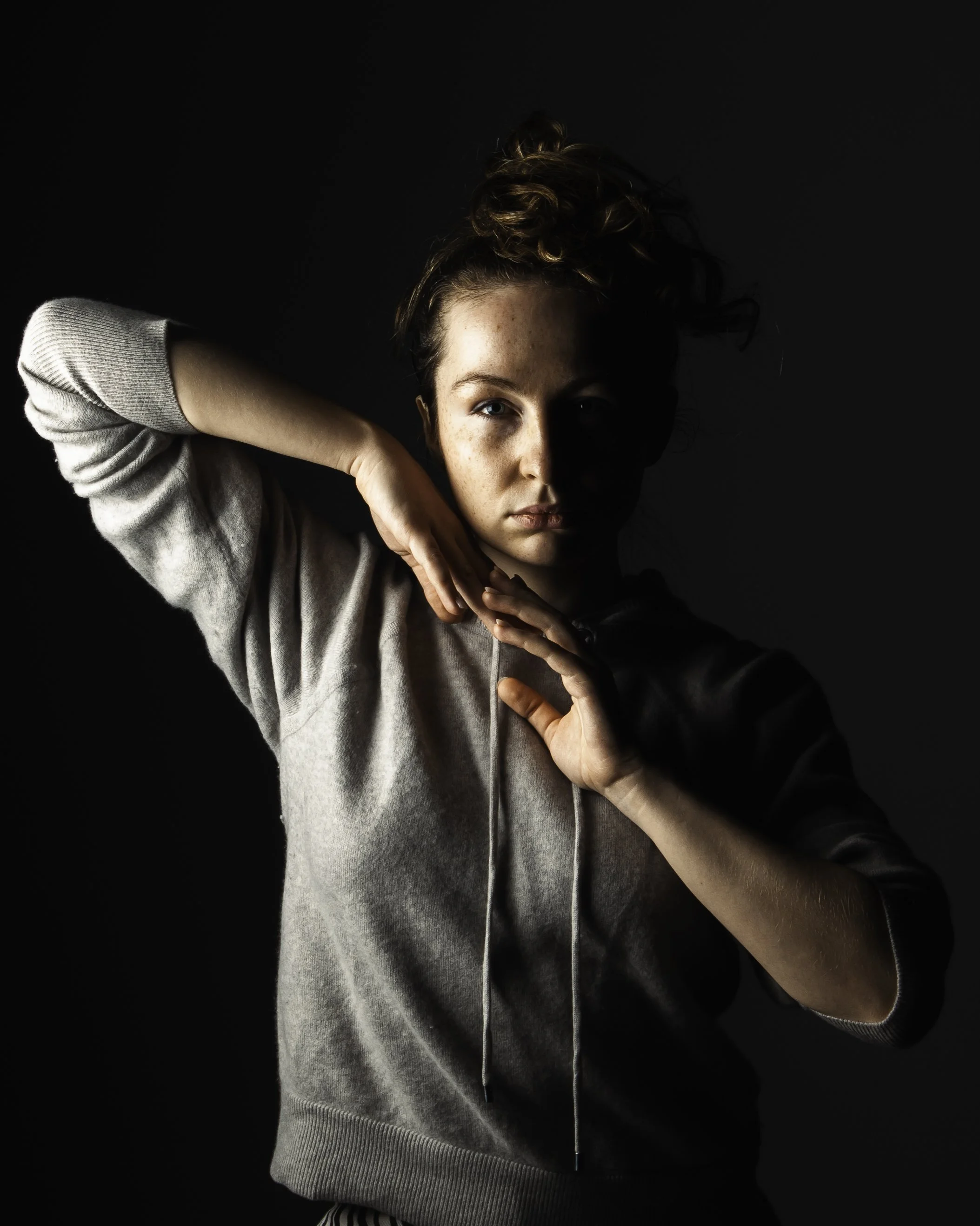A young woman with curly hair in a high bun poses against a dark background, with one arm bent over her head and the other in front of her chest, wearing a gray hoodie.