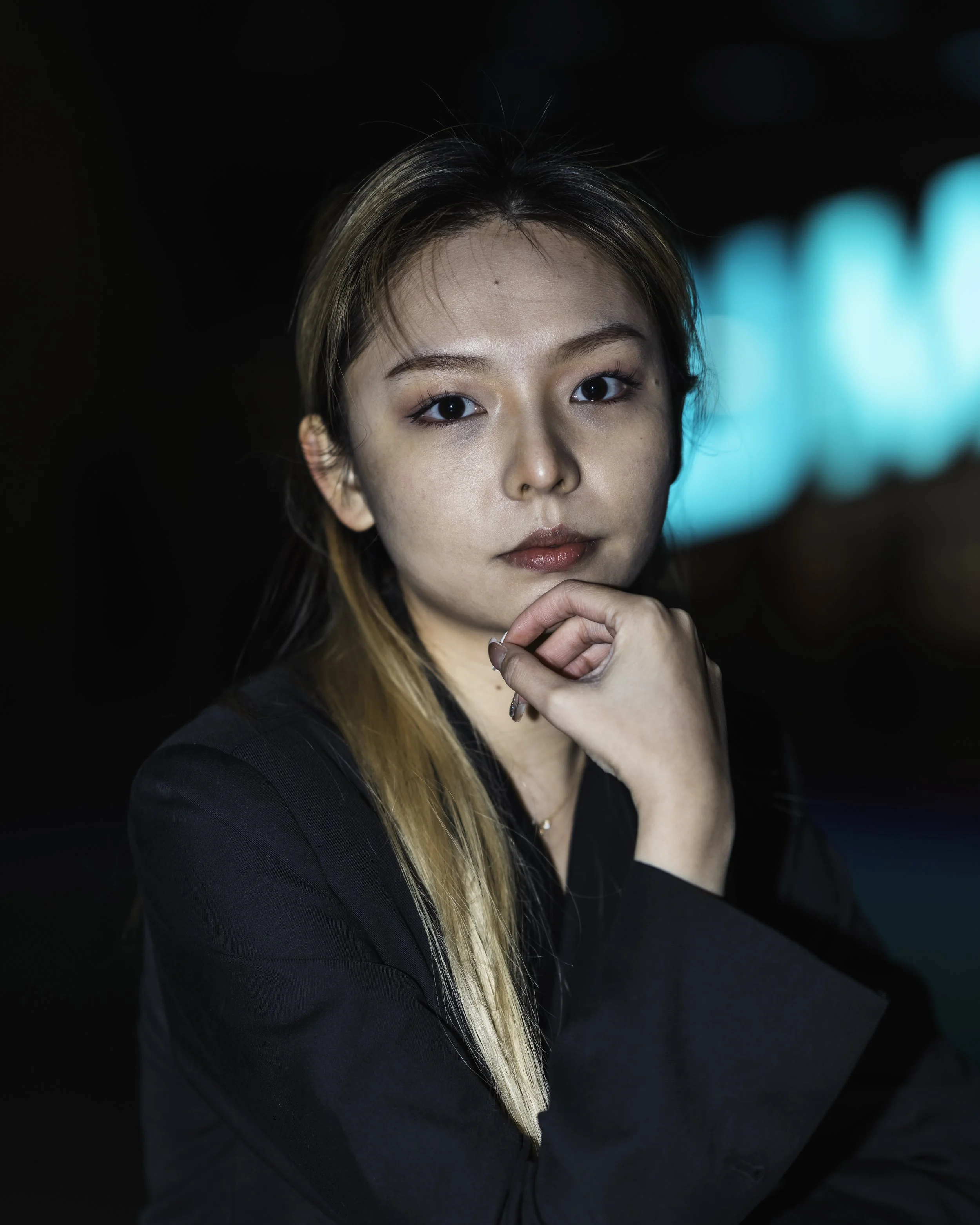 Portrait of a woman with long hair, wearing a dark jacket, looking confidently at the camera with her chin resting on her hand, in a dimly lit setting with a neon blue background.