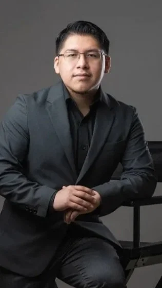 A man in a dark suit and glasses sitting with his hands clasped, looking at the camera.