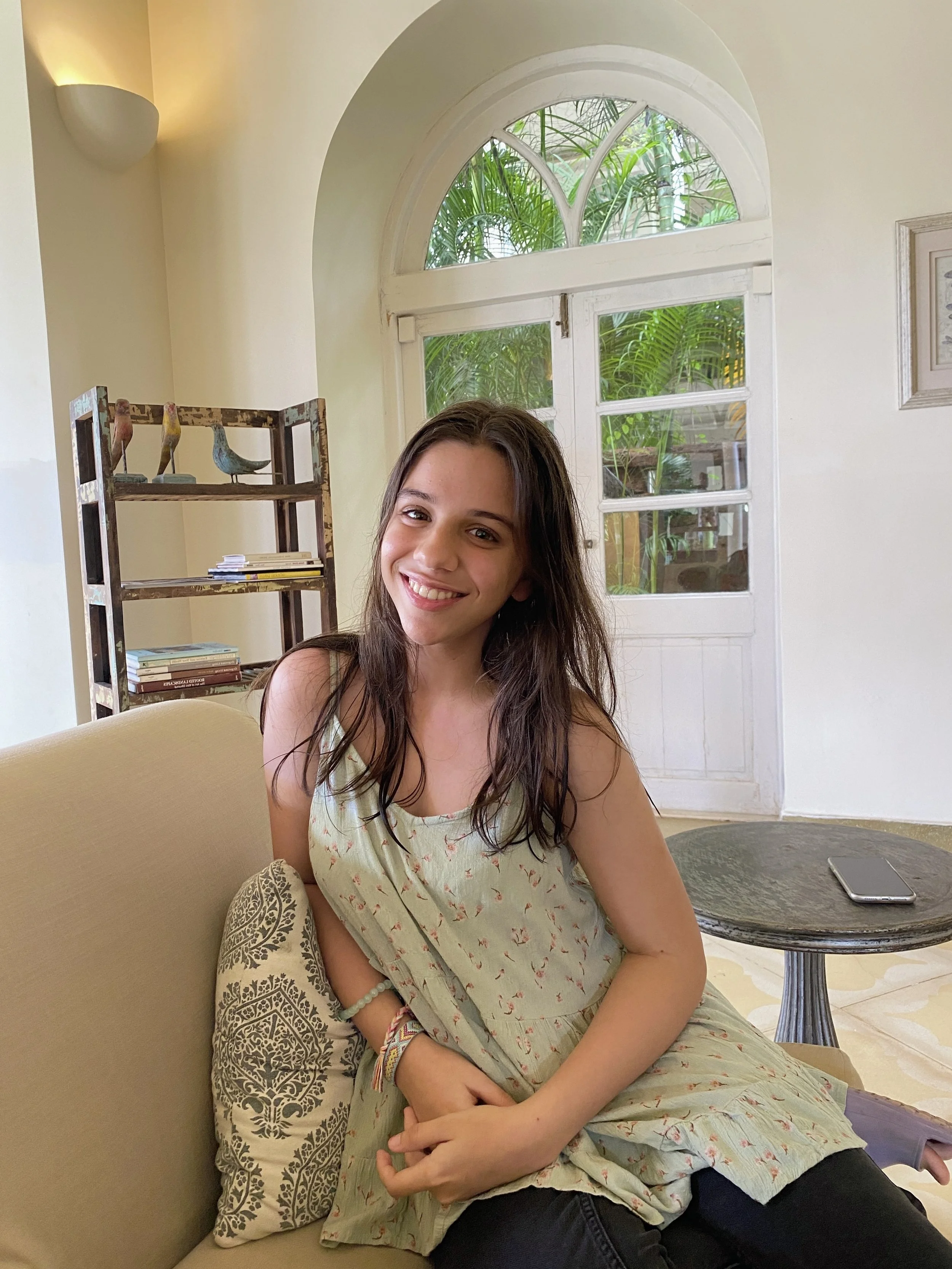 Young woman sitting on a sofa in a cozy room with a smile, green dress, bracelets, and books on a shelf. A round table with a smartphone and a large window with greenery outside are in the background.