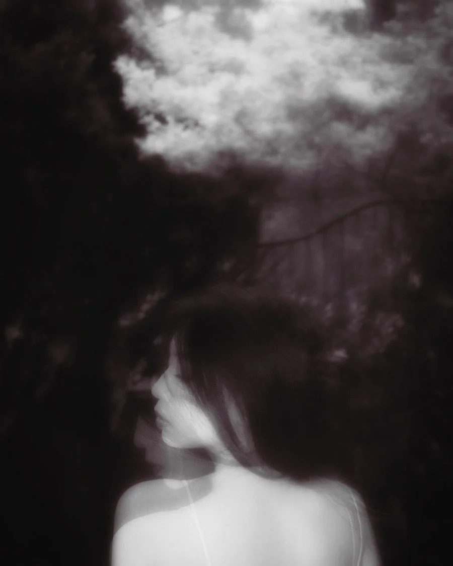 Black and white photograph of a woman with blurred motion effect, looking sideways with a tree or cloud background.