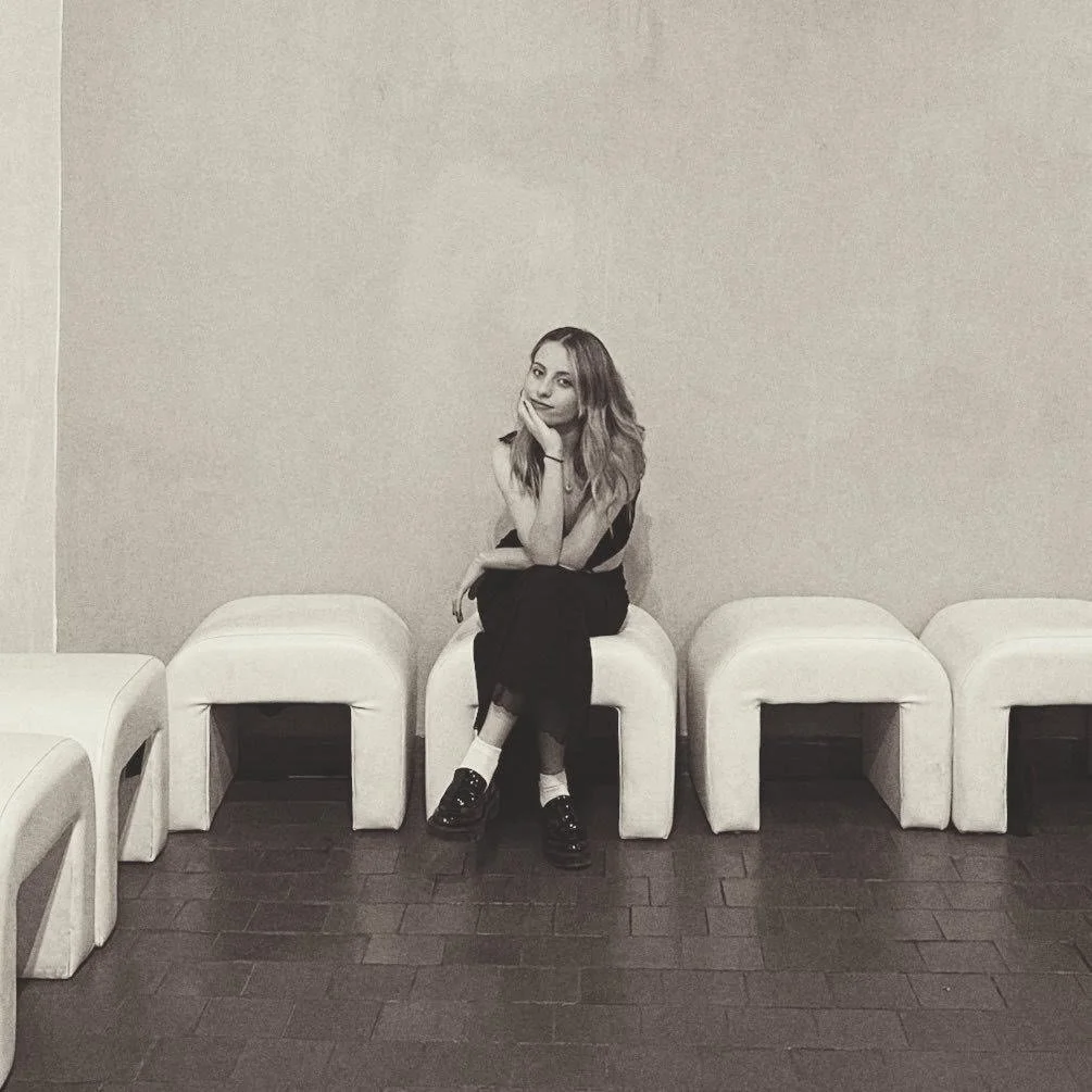 Black and white photo of a woman sitting on a padded bench, surrounded by cushioned stools along a tiled floor against a plain wall.