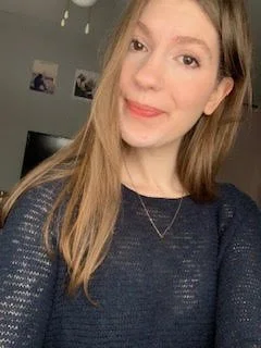 Young woman smiling, long hair, wearing dark sweater, necklace visible, indoor background with blurred wall art and TV.