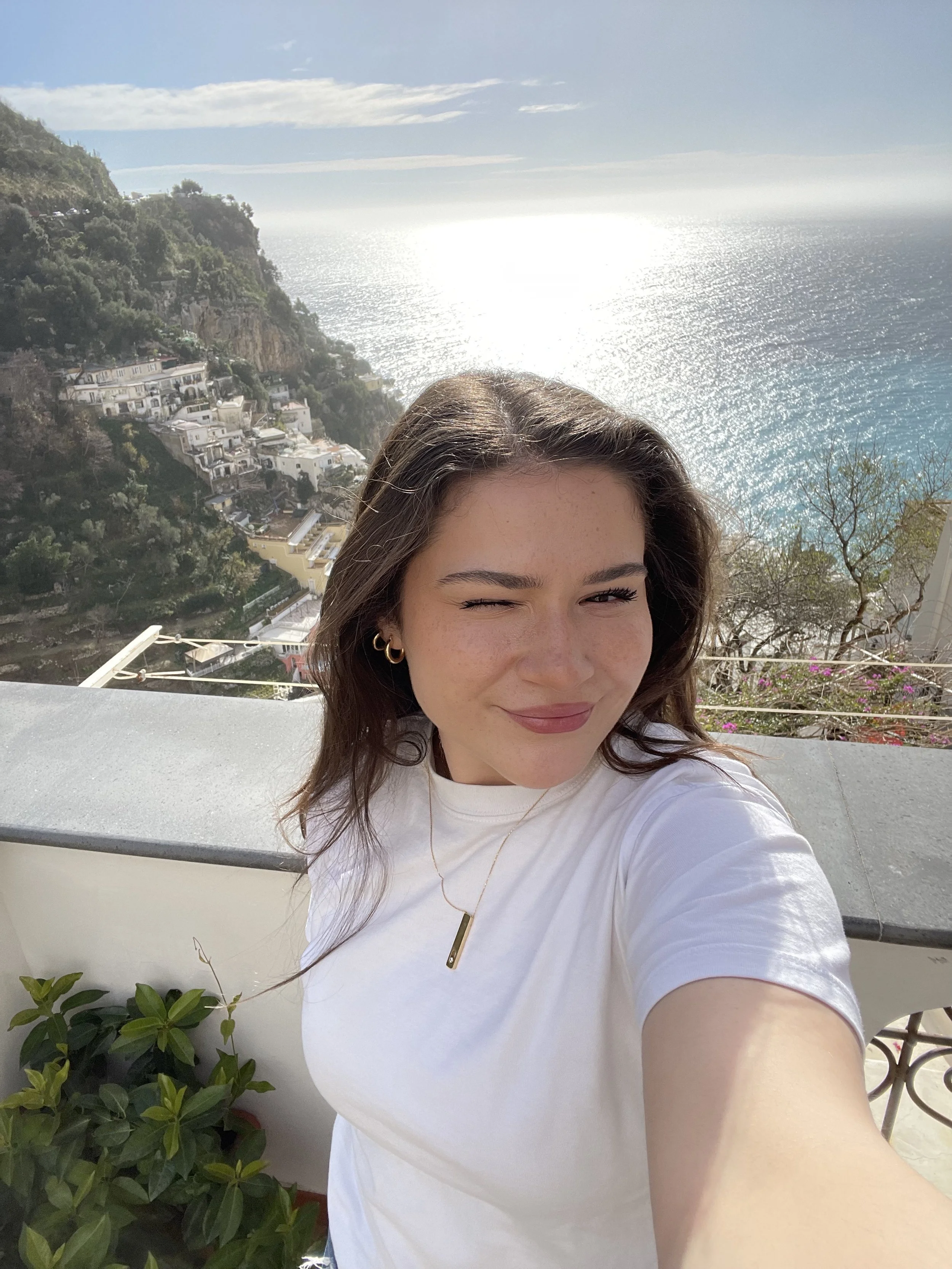 Woman taking a selfie with a scenic ocean view and hillside buildings in the background.