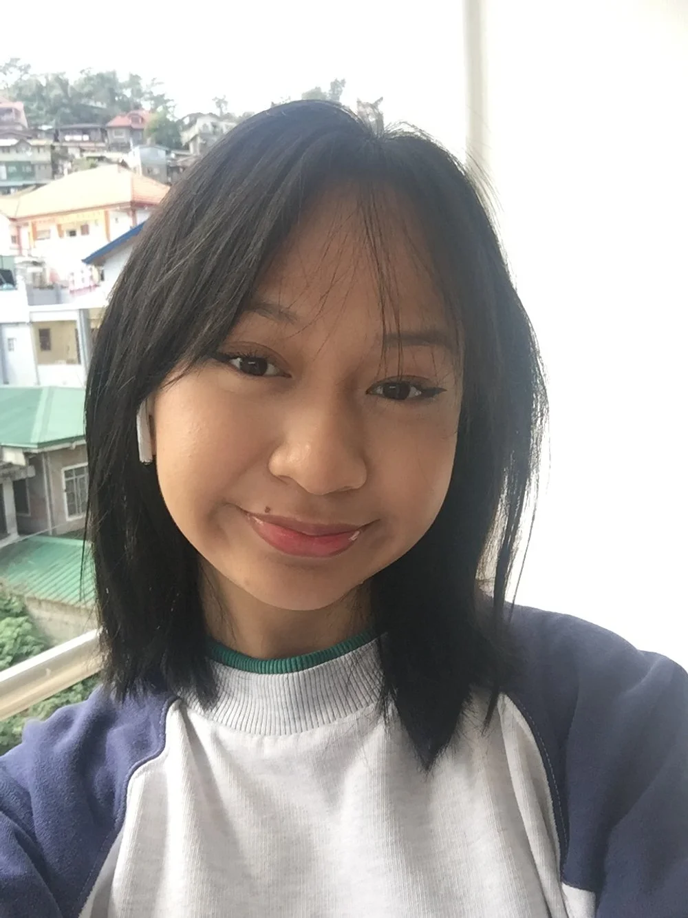 Person with dark hair smiling, wearing white and dark blue top with earbuds, cityscape background.