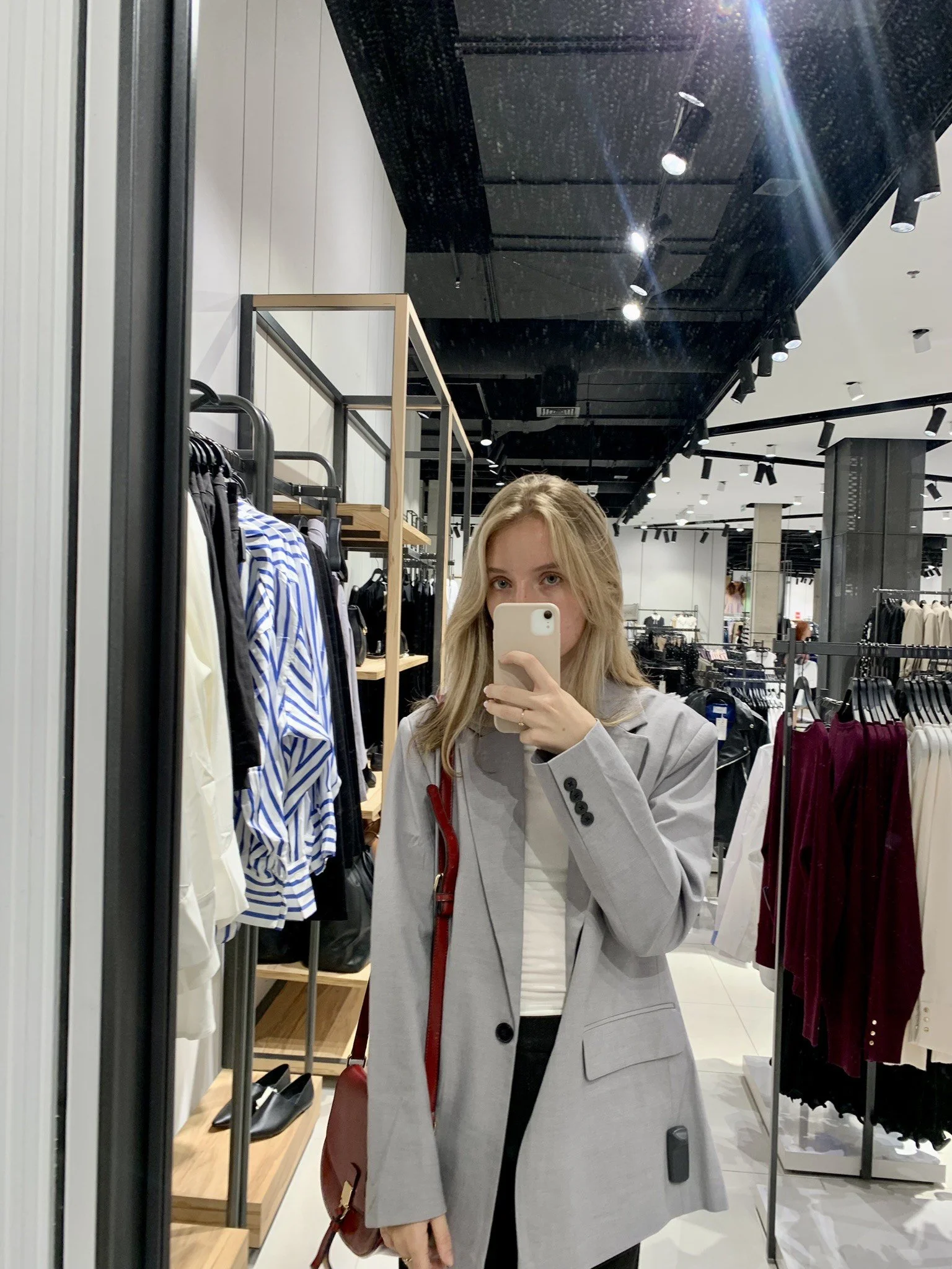Person taking a mirror selfie in a clothing store, wearing a gray blazer and a white shirt, with racks of clothes in the background.