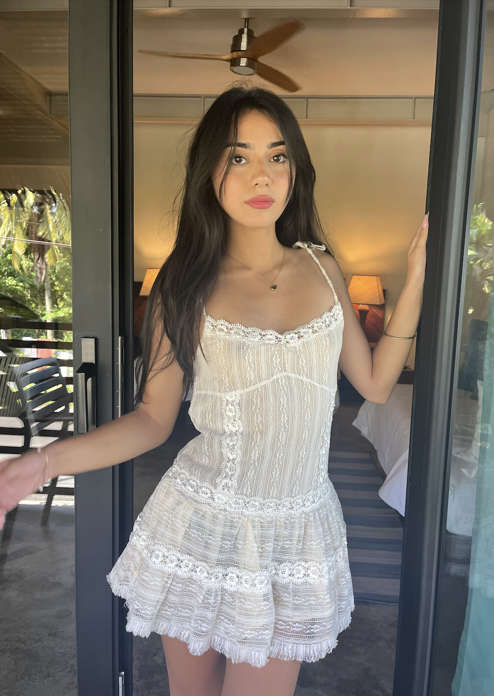 Woman in white lace dress standing at glass doors with an indoor background and a ceiling fan visible.