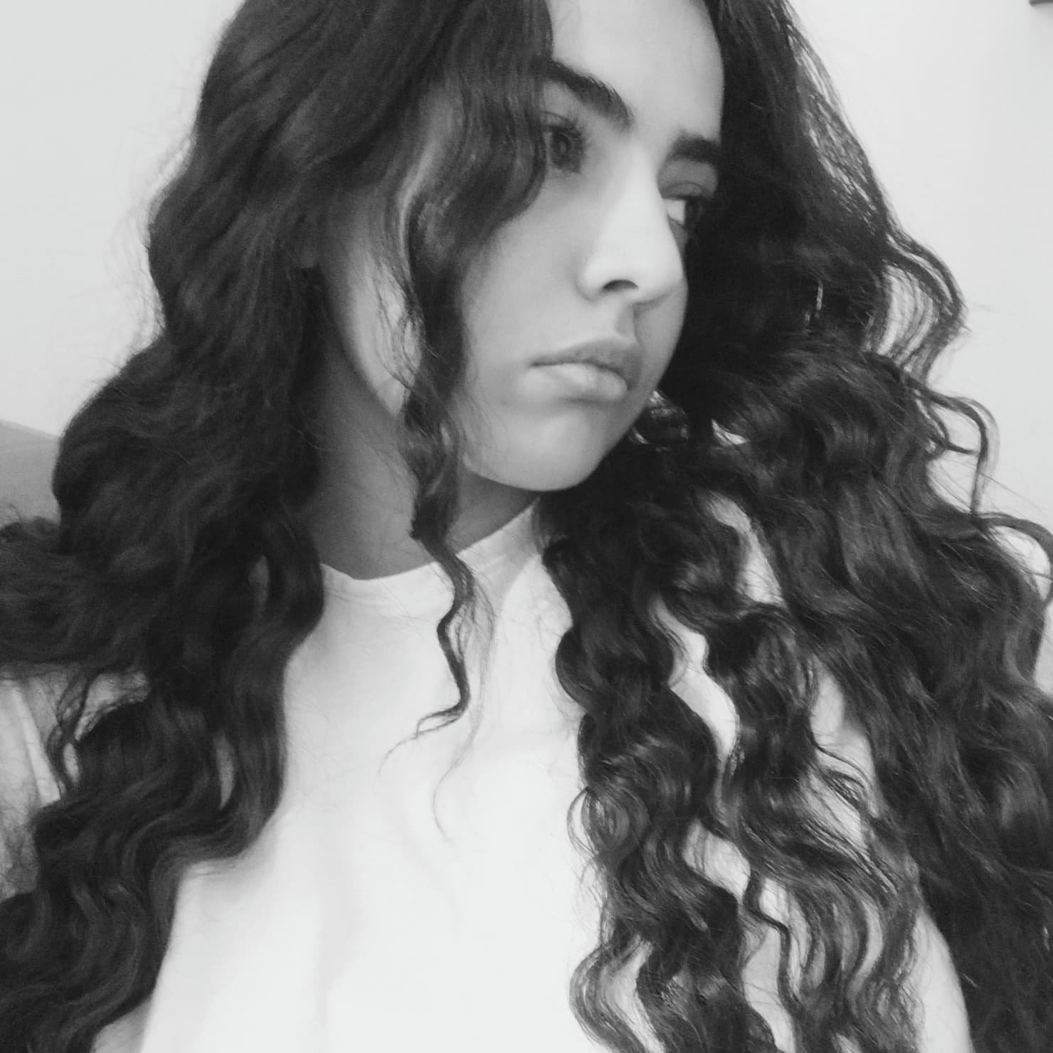Black and white photo of a person with long, curly hair and a neutral expression, wearing a light-colored shirt.