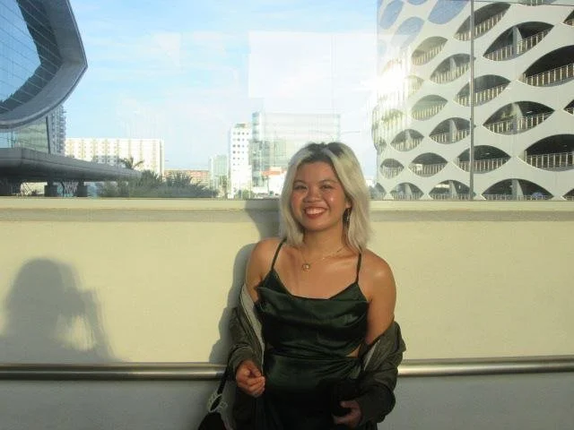 Woman in green dress smiling, standing in front of modern buildings with unique architecture and large windows.