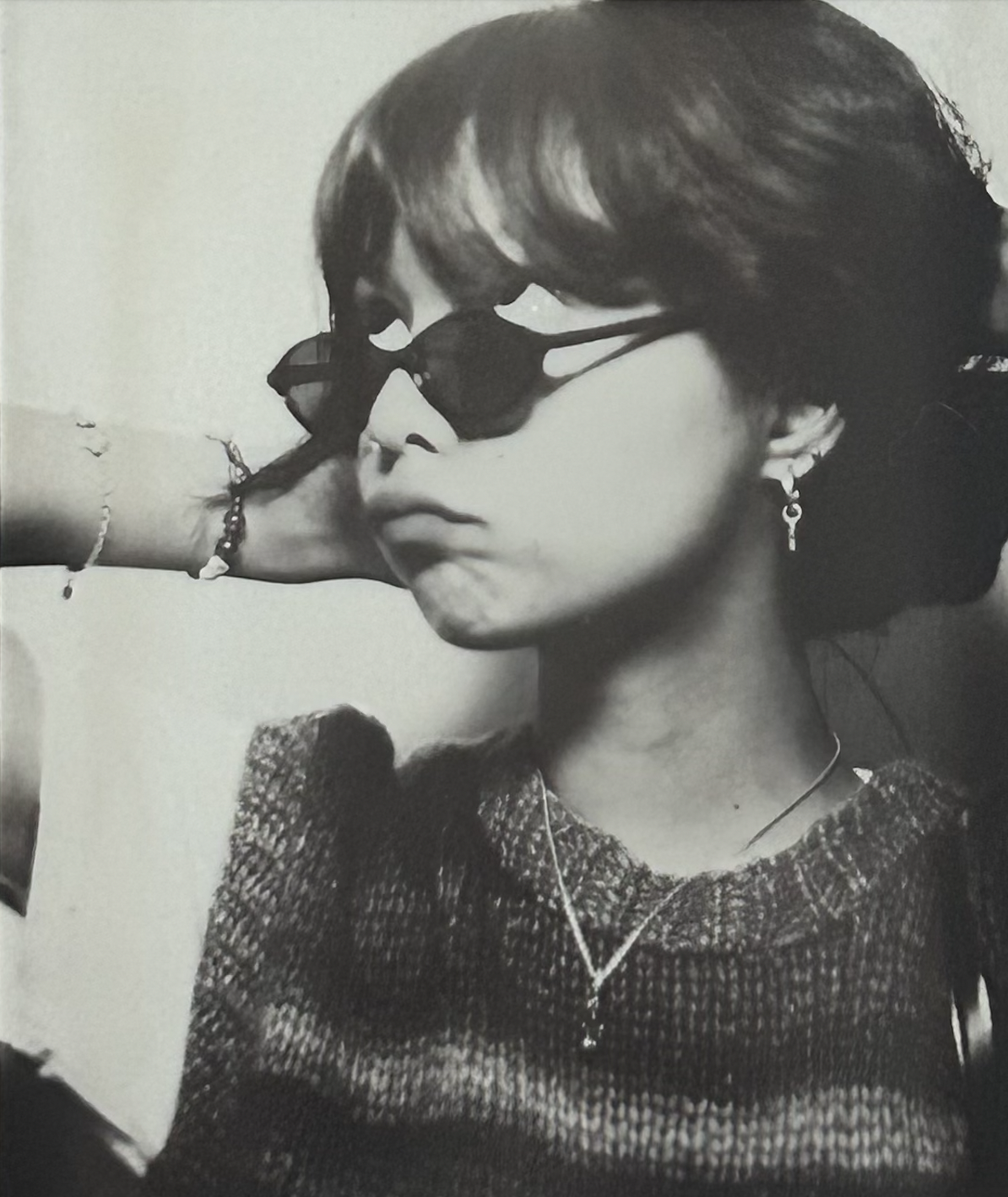 Black and white photo of a young person wearing sunglasses and a knitted sweater, posing with a hand behind their head and displaying a casual expression. They are accessorized with earrings and a necklace.