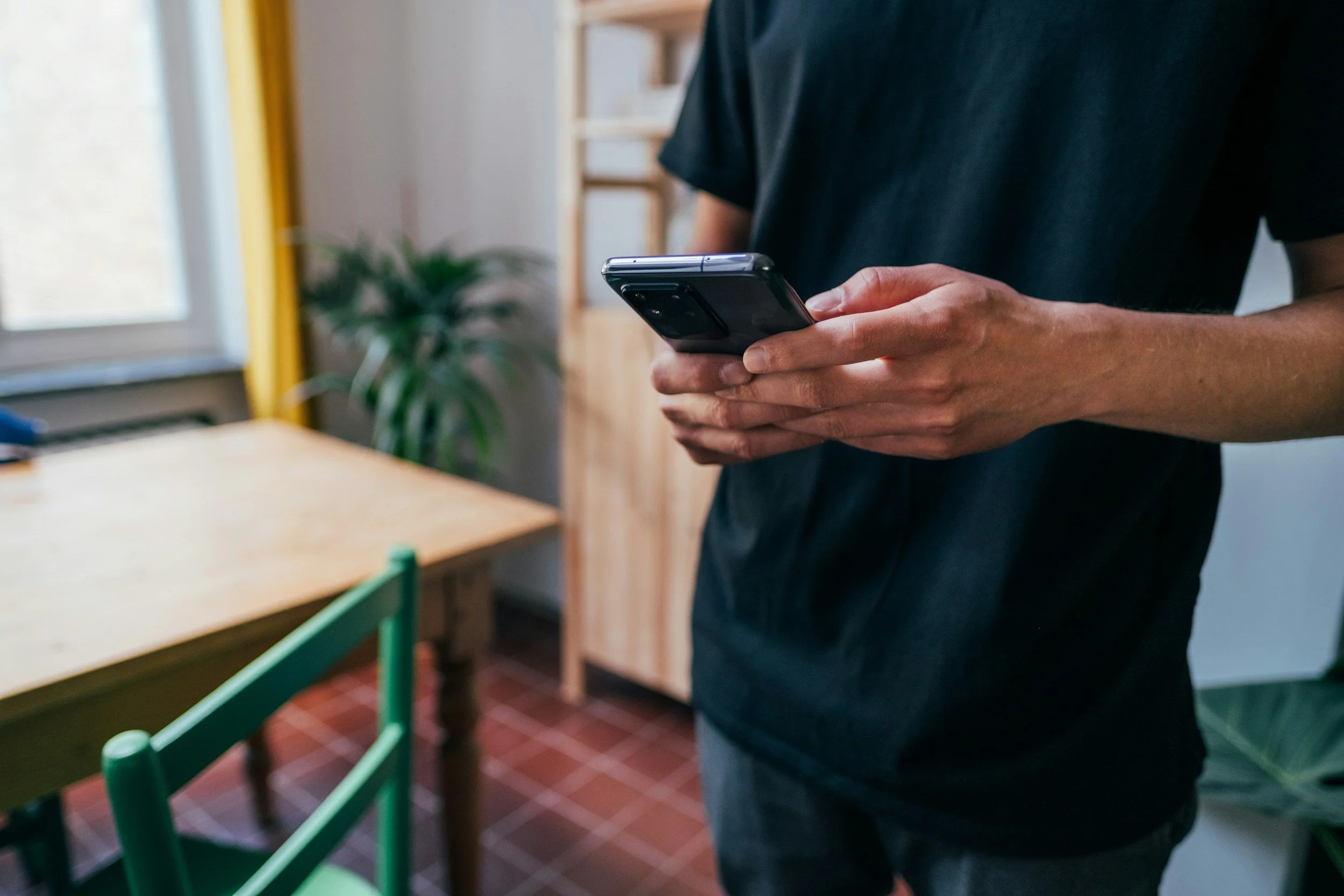 Person in black shirt holding a smartphone indoors near a green chair, window, and wooden furniture.