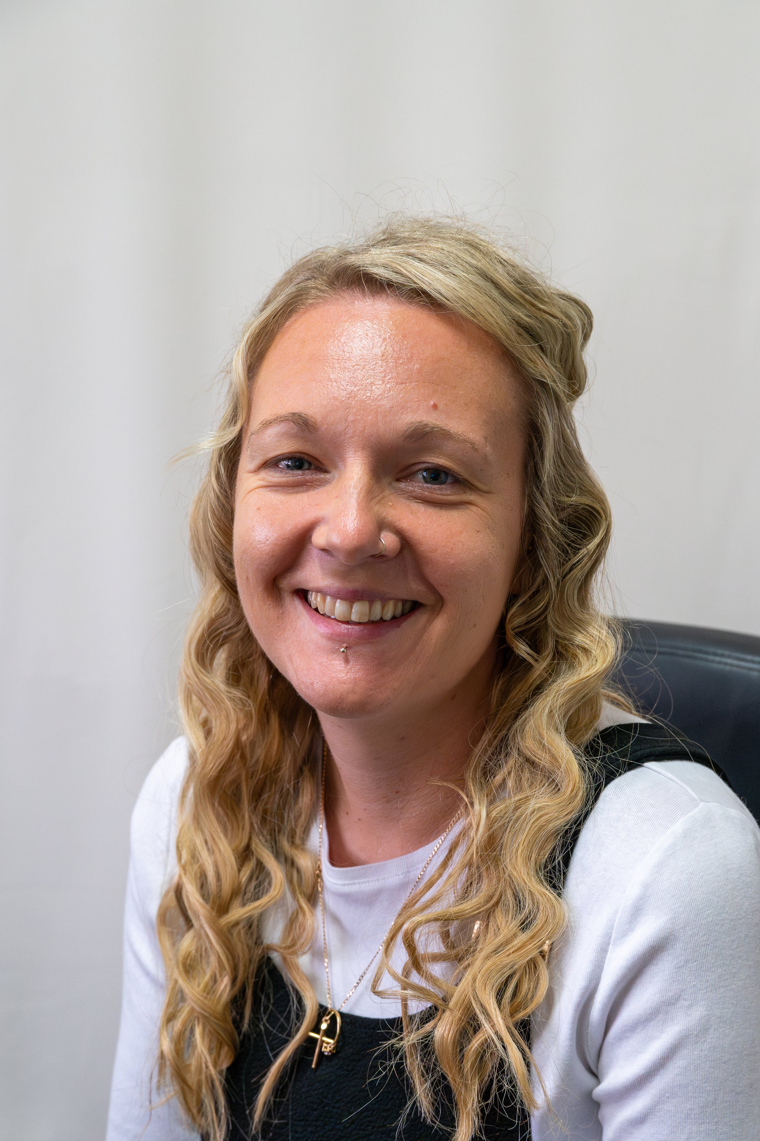 A smiling woman with long blonde hair, wearing a white shirt, hoop earrings, and a necklace with a key pendant, standing indoors against a plain white wall.