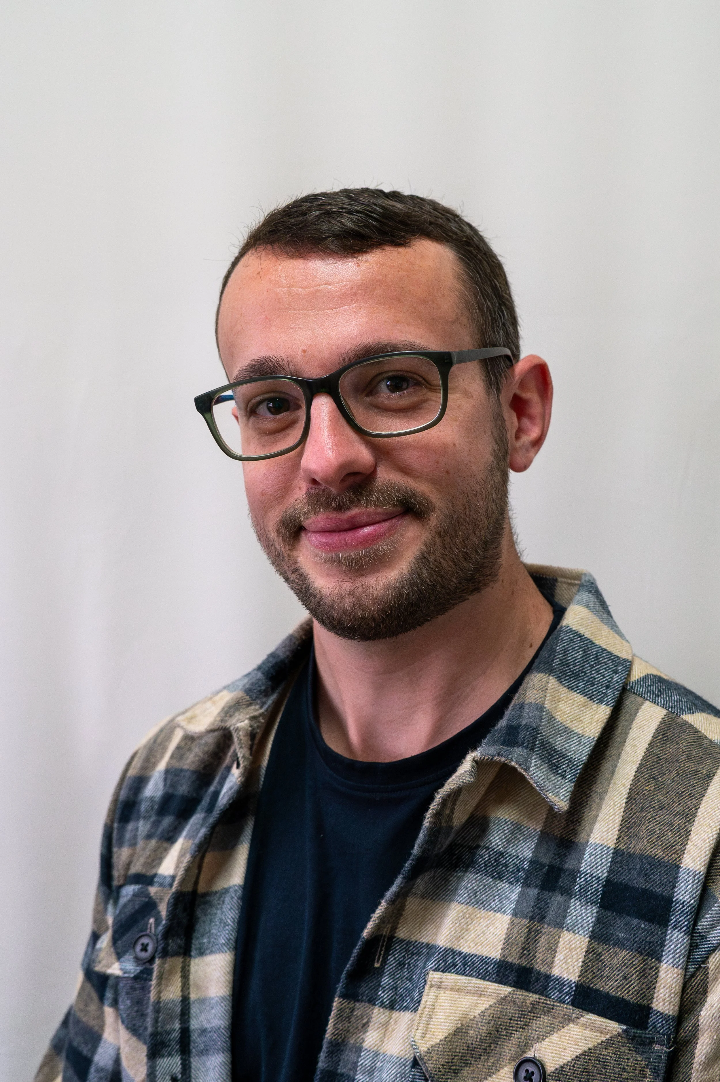 A young man with short dark hair, glasses, and a beard, wearing a light blue denim button-up shirt, standing against a plain light-colored background.