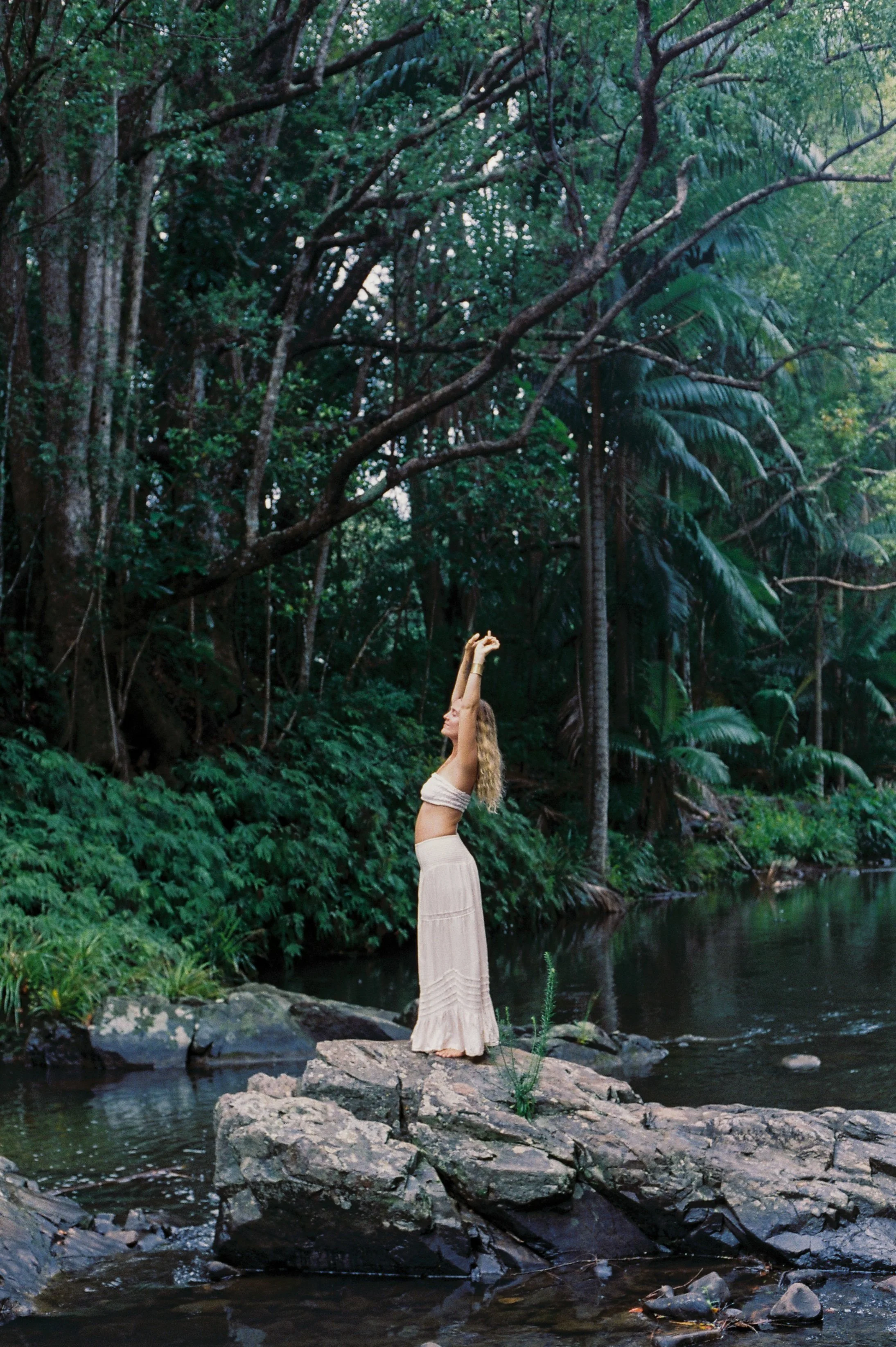 A woman standing on large rocks in a river, stretching with arms raised, surrounded by dense green foliage and trees.