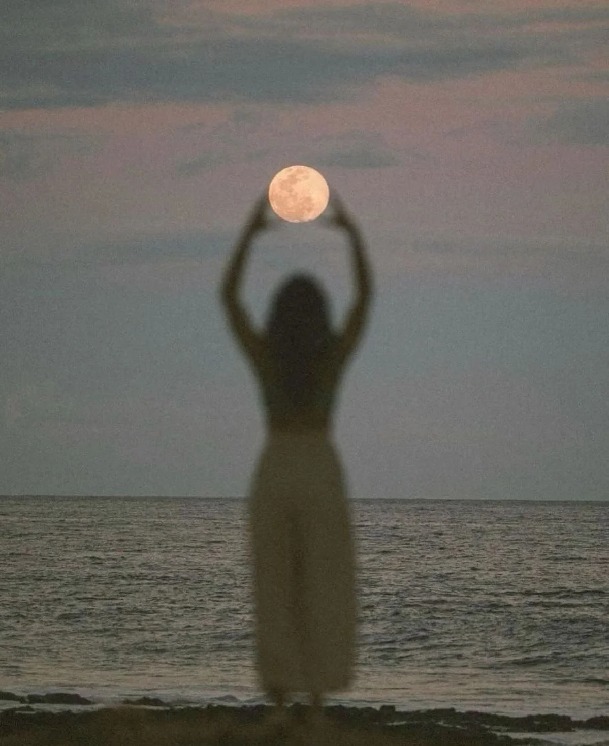 Silhouette of a woman on a beach at sunset, holding her hands above her head, creating a frame with her arms around the full moon in the sky.