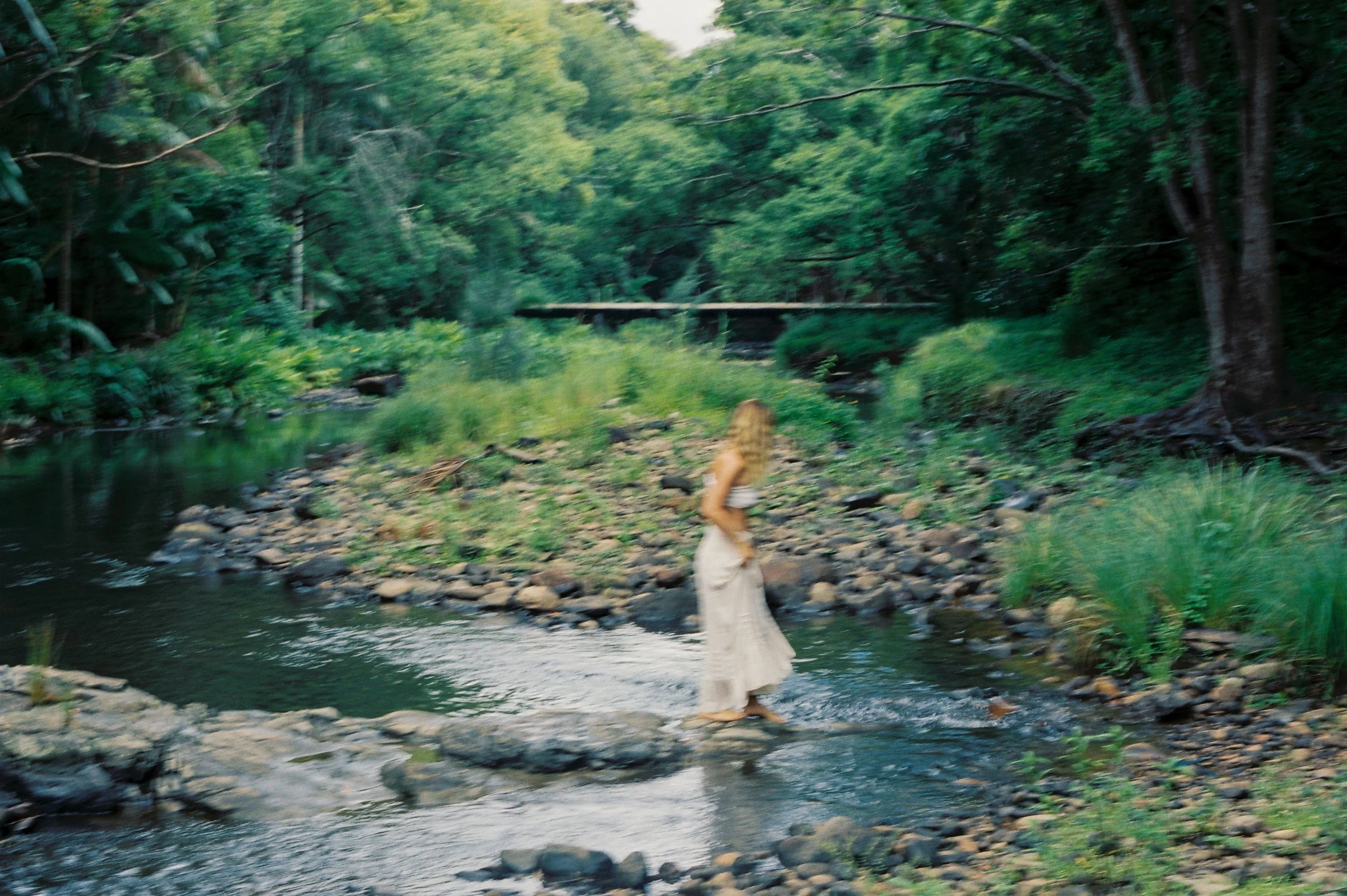 A woman crossing a small stream in a lush green forest.