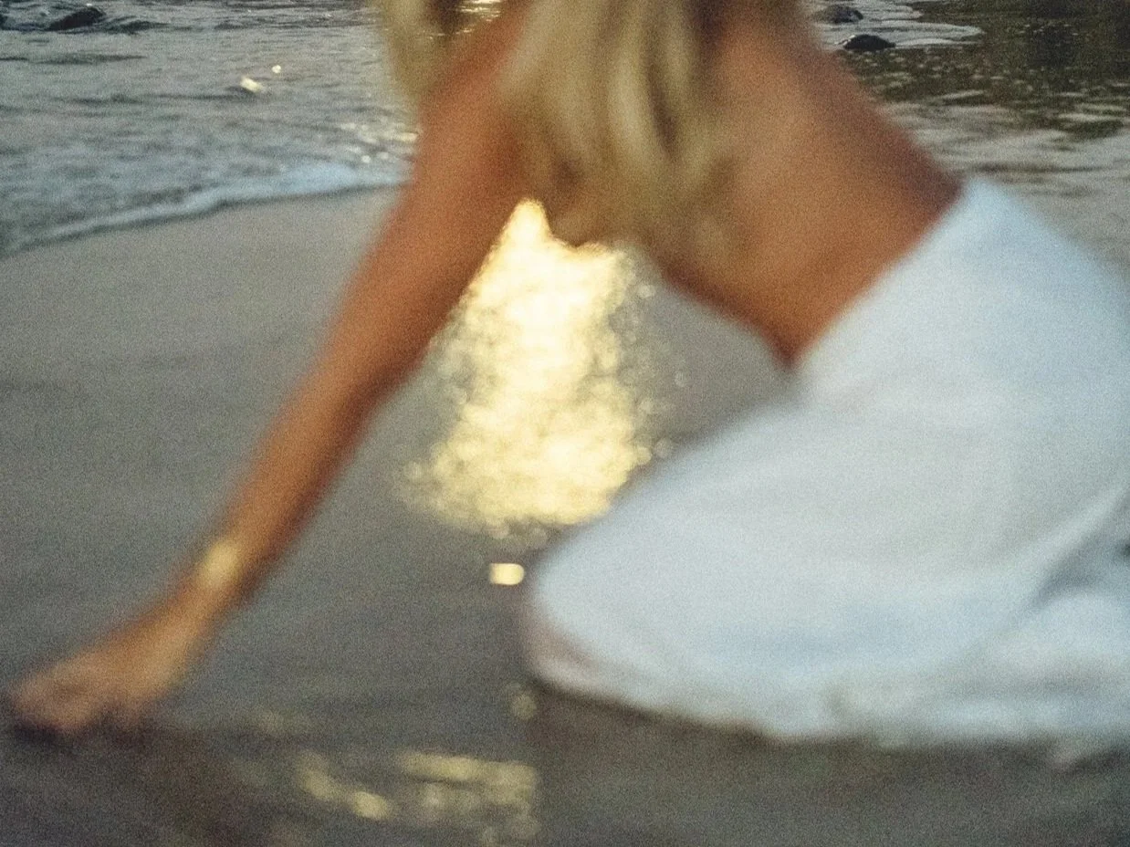 A woman with blonde hair wearing a white dress is kneeling on the wet sand at the beach, with the ocean in the background.